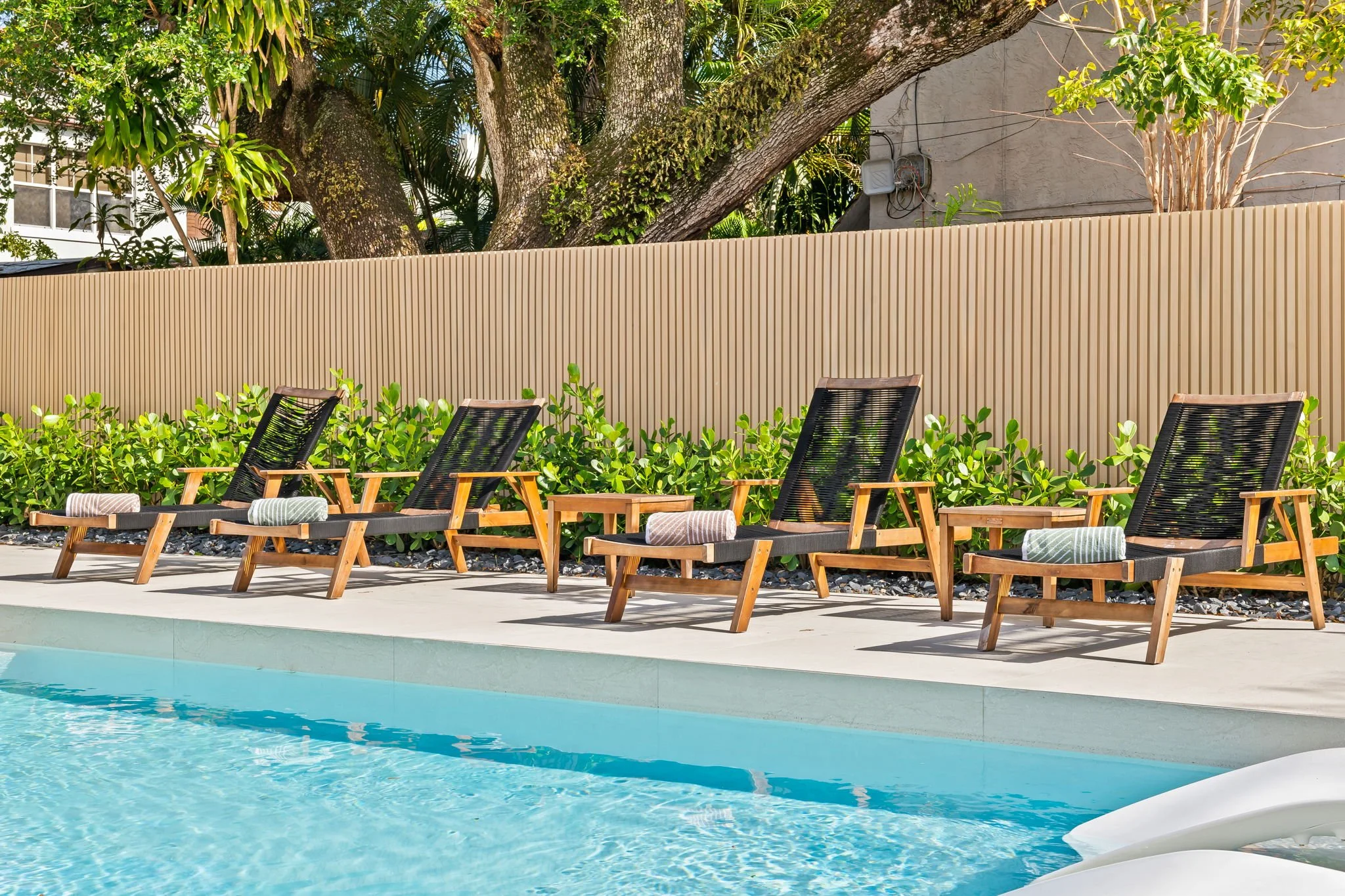 view of a swimming poolside with multiple black lounge chairs with striped cushions, surrounded by lush green plants, trees, and a beige wooden fence.