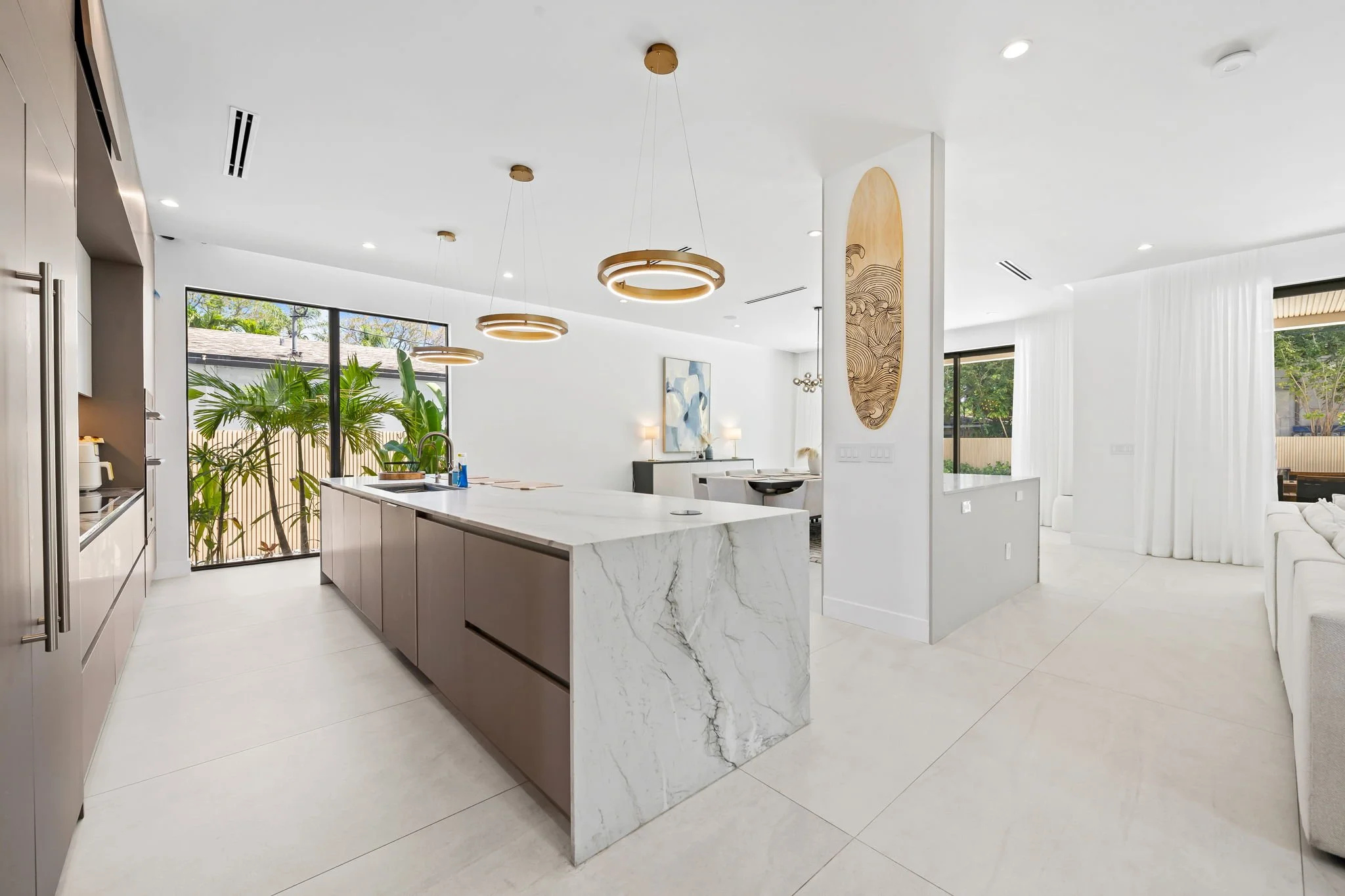 Modern open-plan kitchen and living space with a large white marble island, pendant lighting, and large windows with view of outdoors.