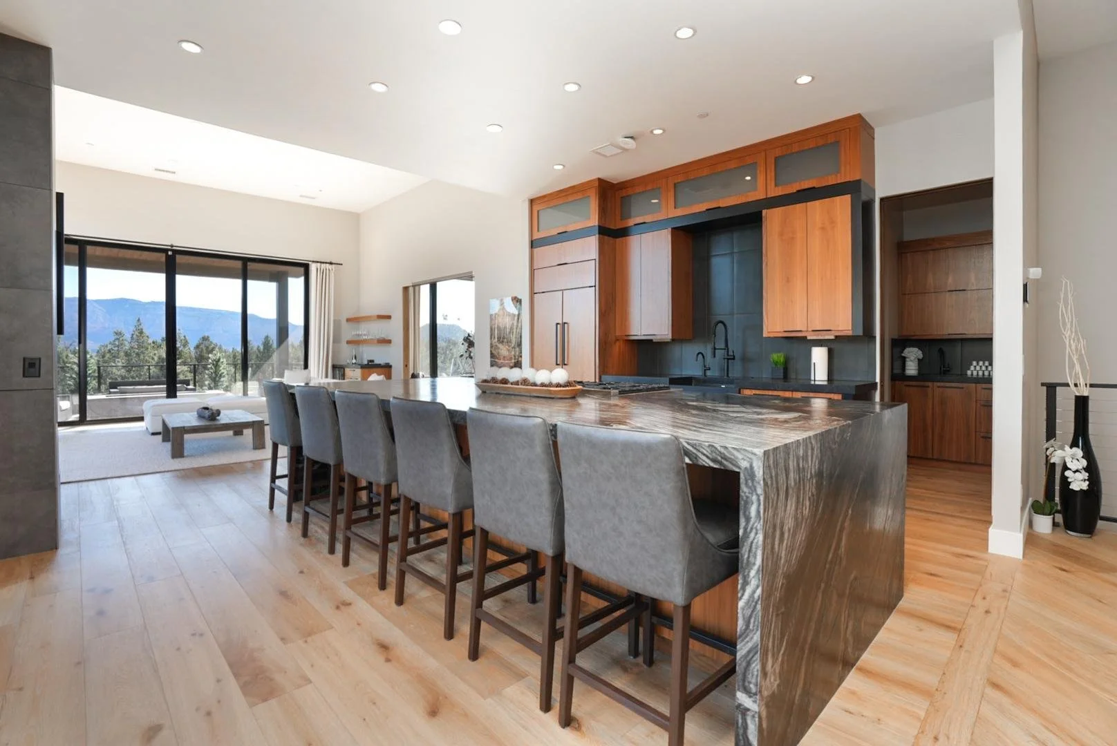 Modern open-concept kitchen with wooden cabinetry, a marble island with bar stools, and a living space with large sliding glass doors leading to a mountain view