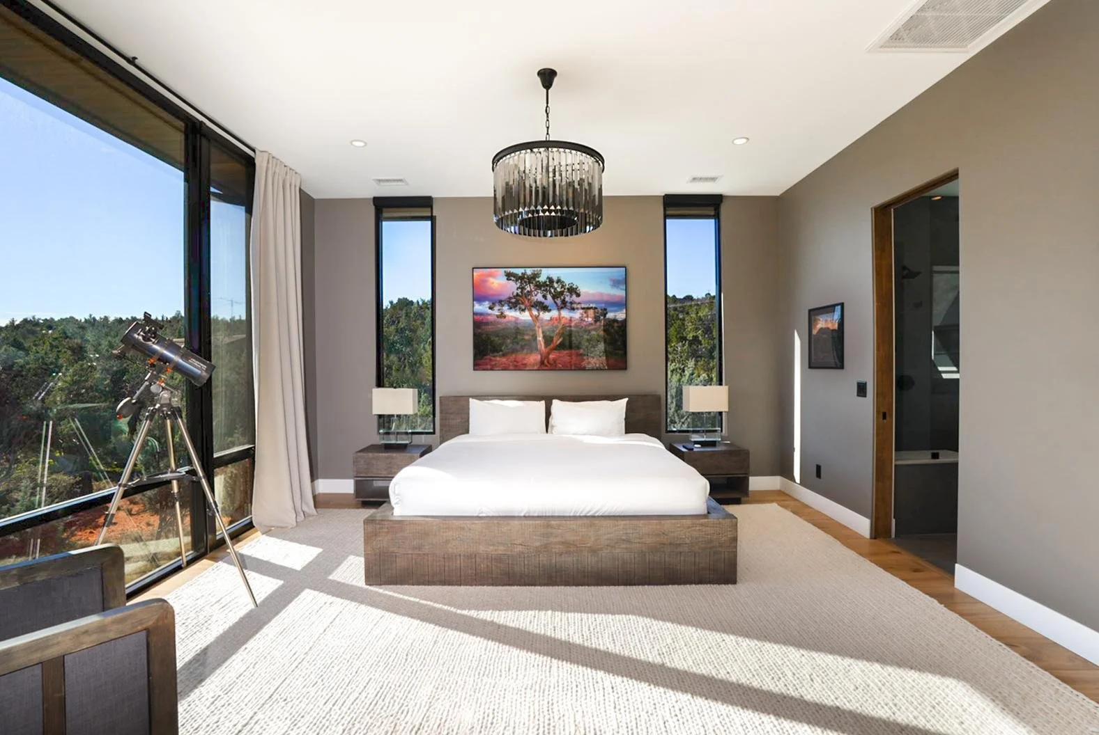 Modern bedroom with a large bed, two nightstands with lamps, a landscape painting above the bed, a telescope near the window, and a chandelier hanging from the ceiling.