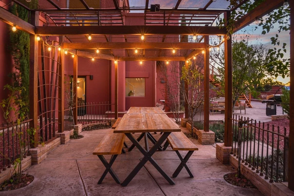 Outdoor patio area with wooden picnic table under a pergola decorated with string lights, surrounded by plants and a black metal fence, with a pink building in the background during sunset.