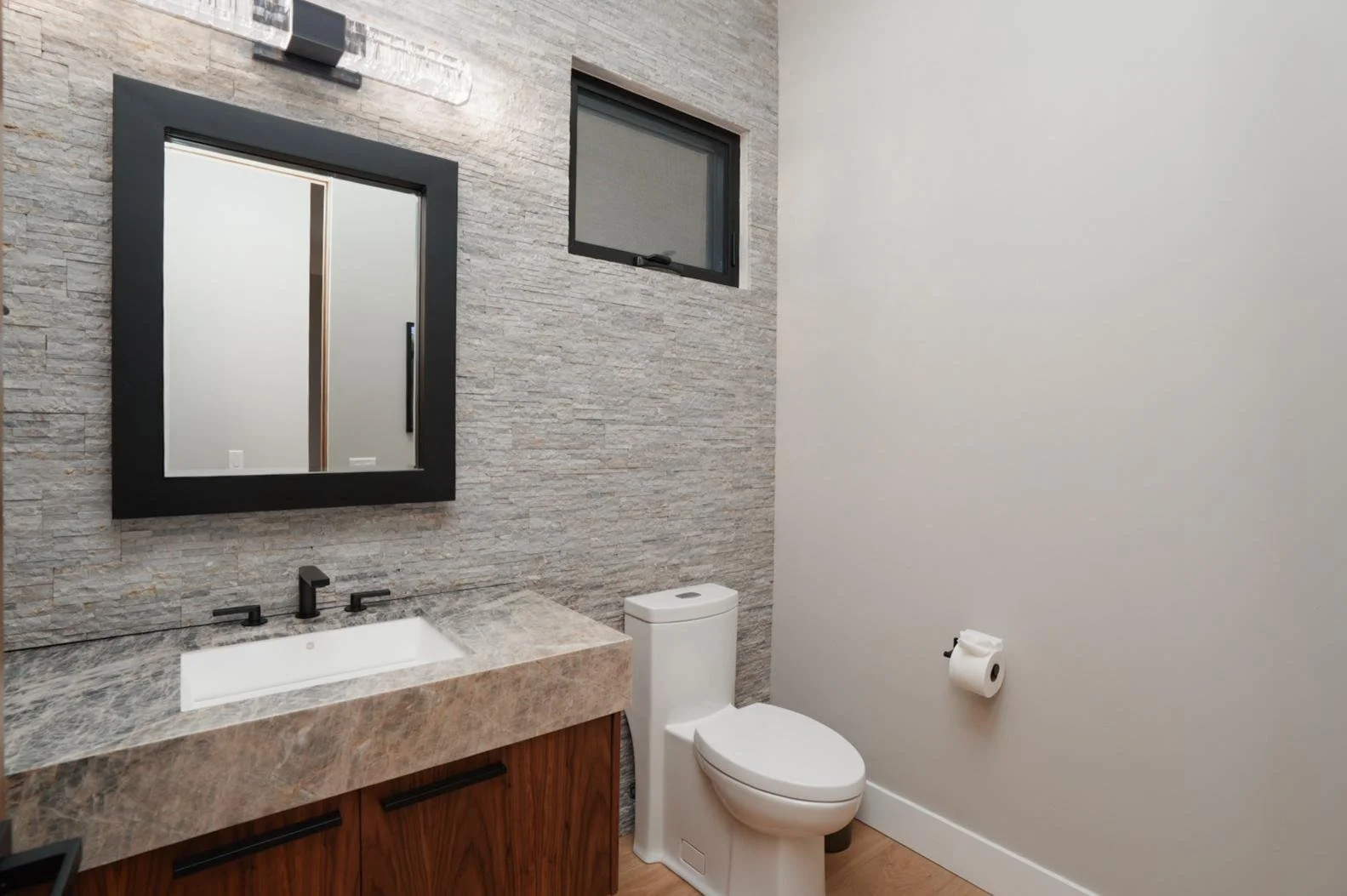 A modern bathroom with a stone wall, a black-framed mirror, a small window, a white toilet, a marble sink counter, and a toilet paper roll on the wall.