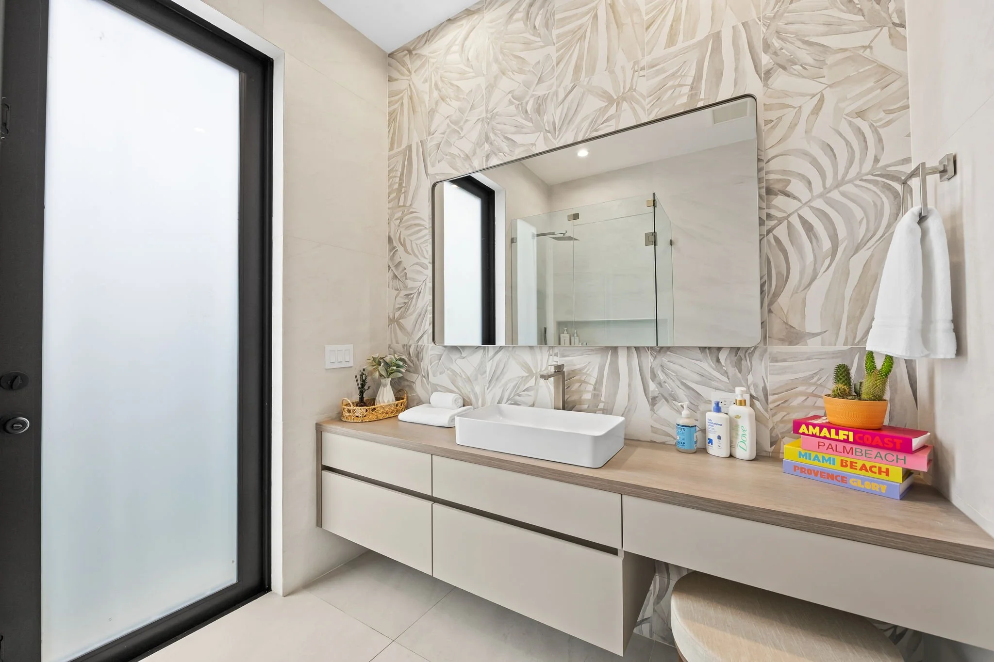 Modern bathroom with a large mirror, decorative wall tiles, a white vessel sink on a wooden vanity, and various toiletries and decorative items, including potted plants and colorful stacked books.