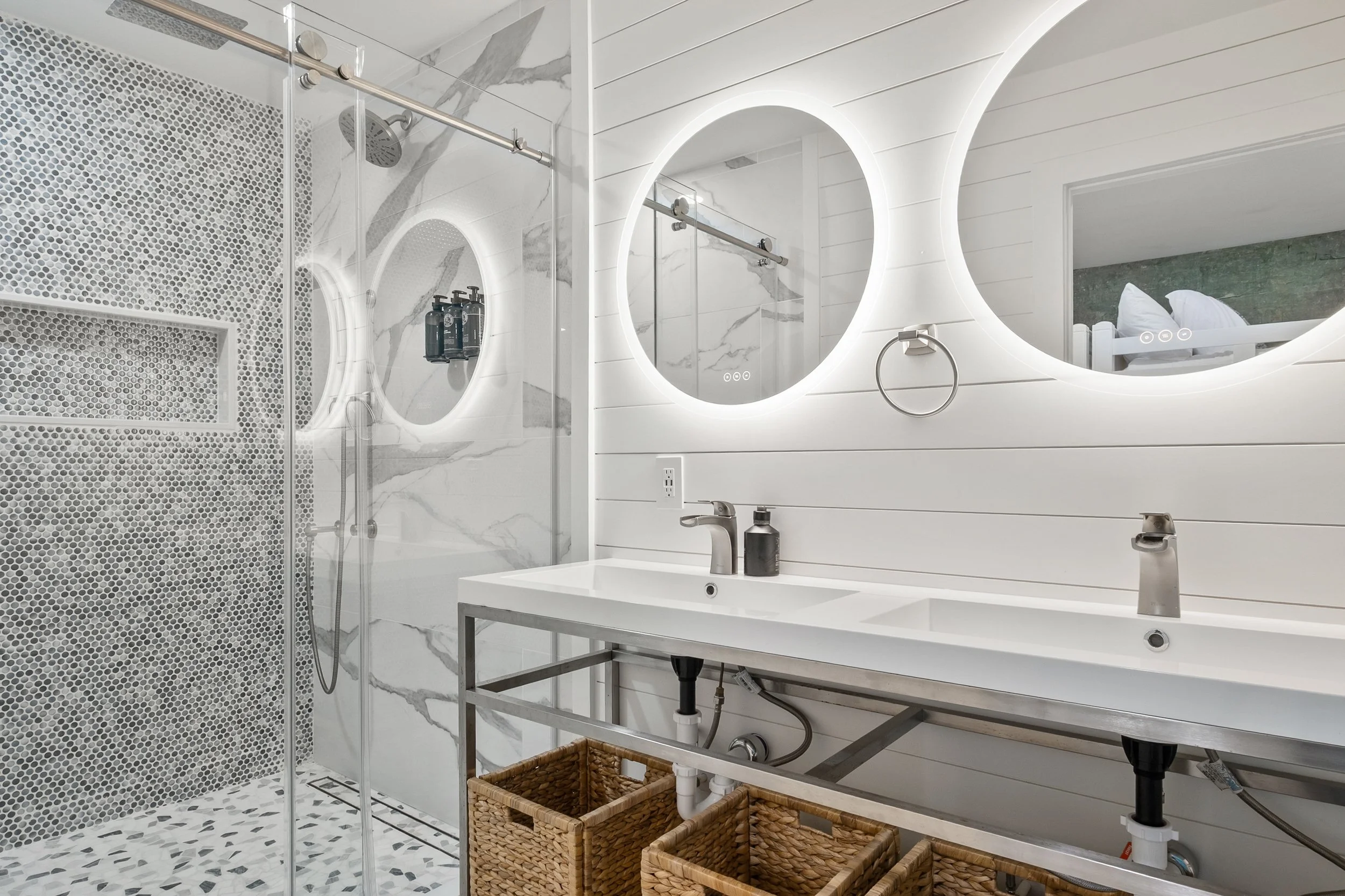 Modern bathroom with double vanity, backlit round mirrors, white shiplap walls, and a walk-in shower with glass door and pebble tiles on the floor.