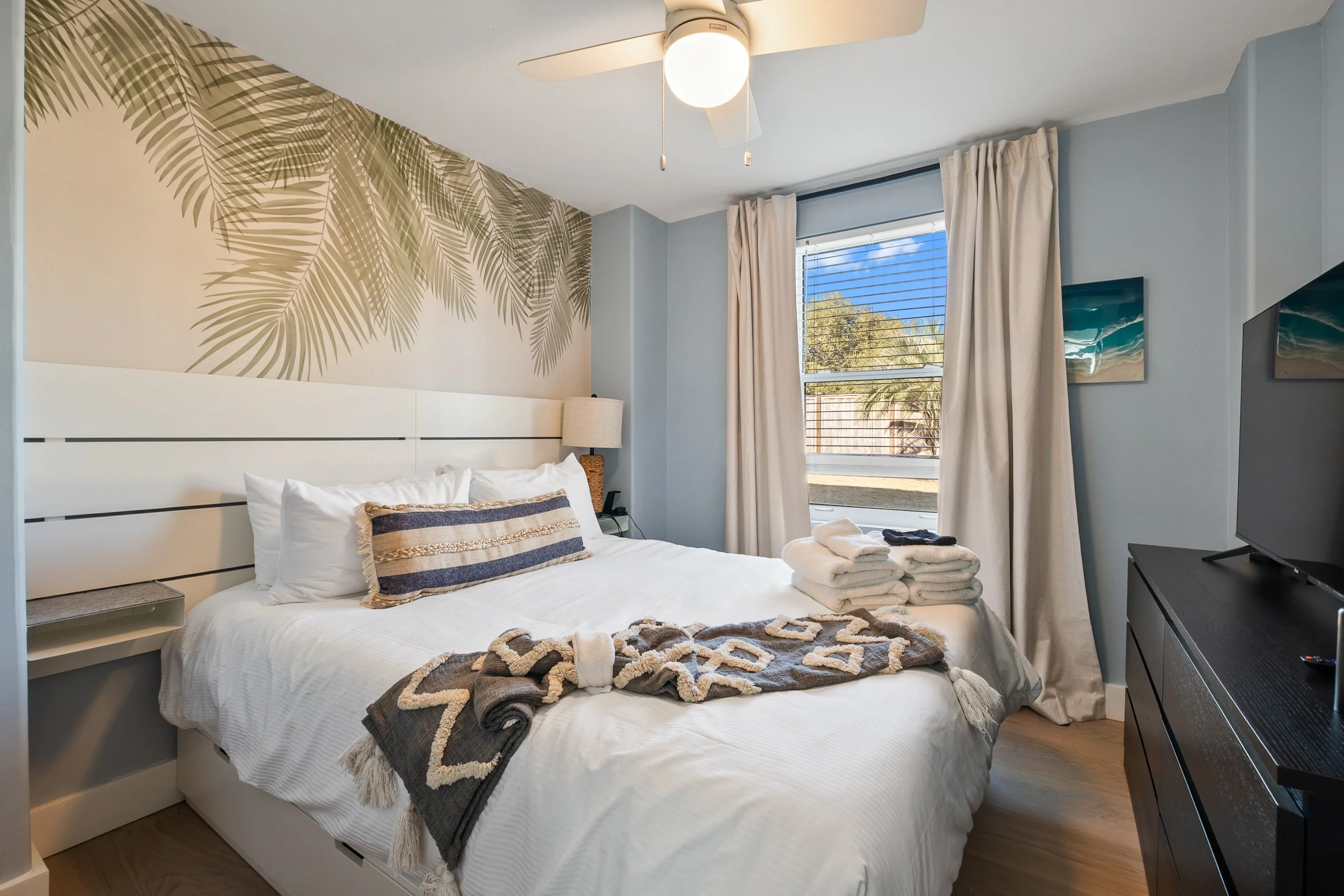 A bedroom with a bed, a window, a dresser with a TV, and decorative wall art of palm leaves. The bed has white linens, a decorative pillow, and towels. There are folded towels and rolled towels at the foot of the bed.