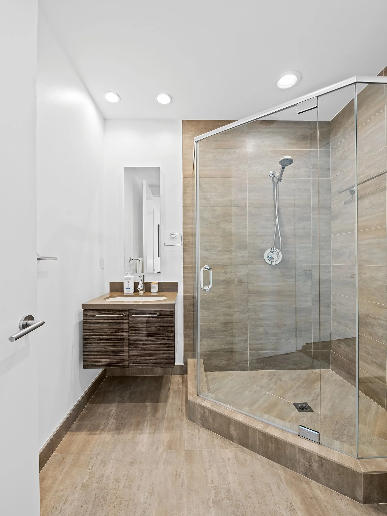 Modern bathroom with glass shower enclosure, beige tile walls and floor, white ceiling, and vanity with a mirror and toiletries.