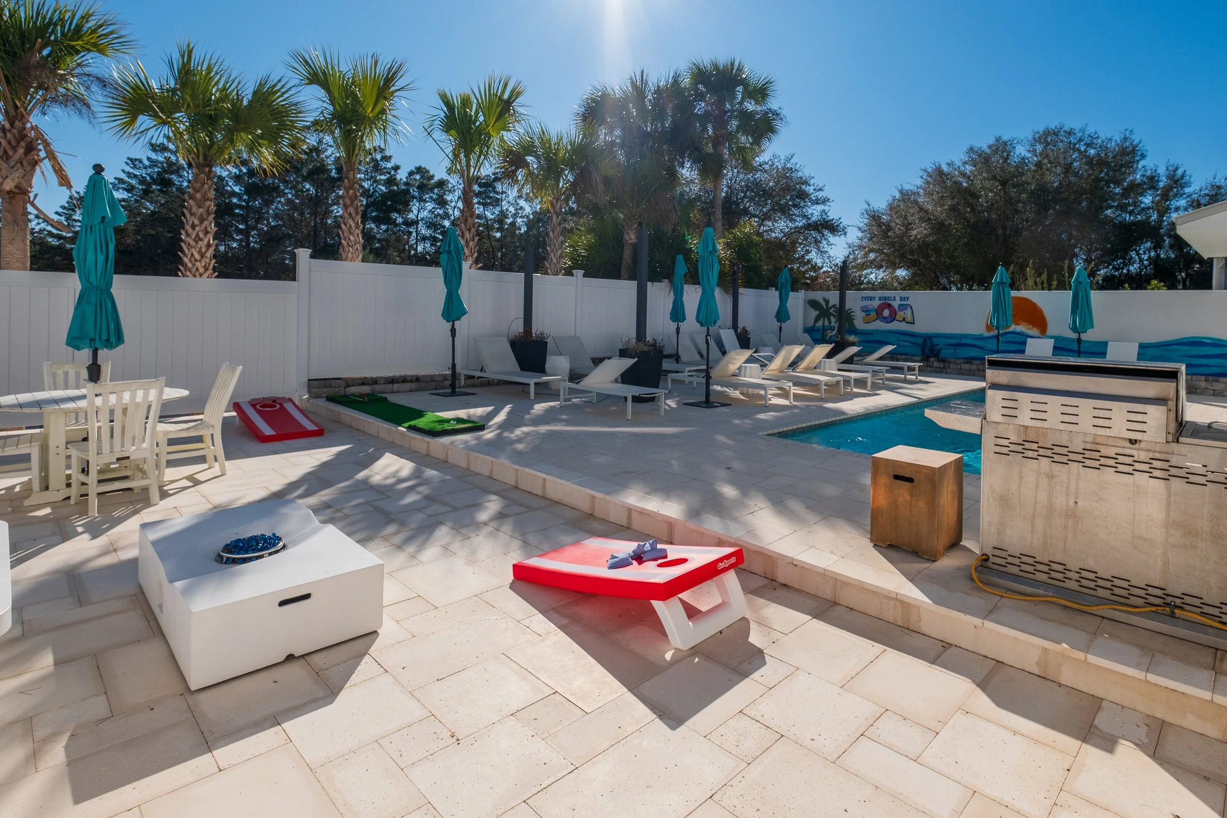 Empty backyard pool area with lounge chairs, umbrellas, a table and chairs, a cornhole game, and a mural on the wall with tropical scene