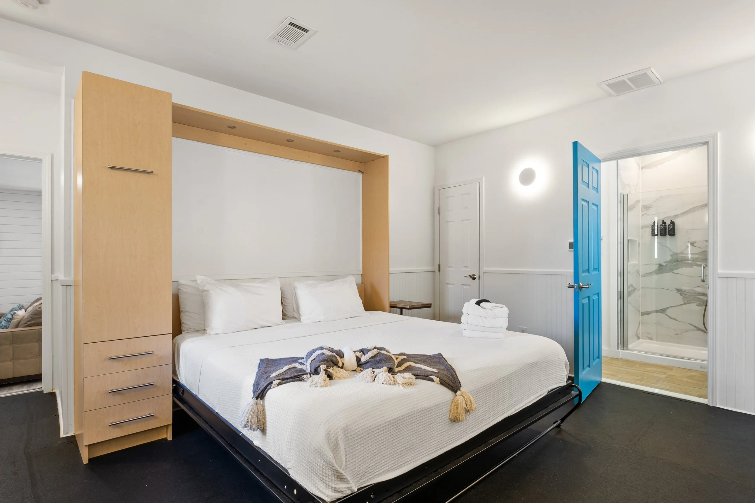 A bedroom with a large bed, white bedding, and a stack of towels, with an en-suite bathroom visible through an open door that has a blue frame.