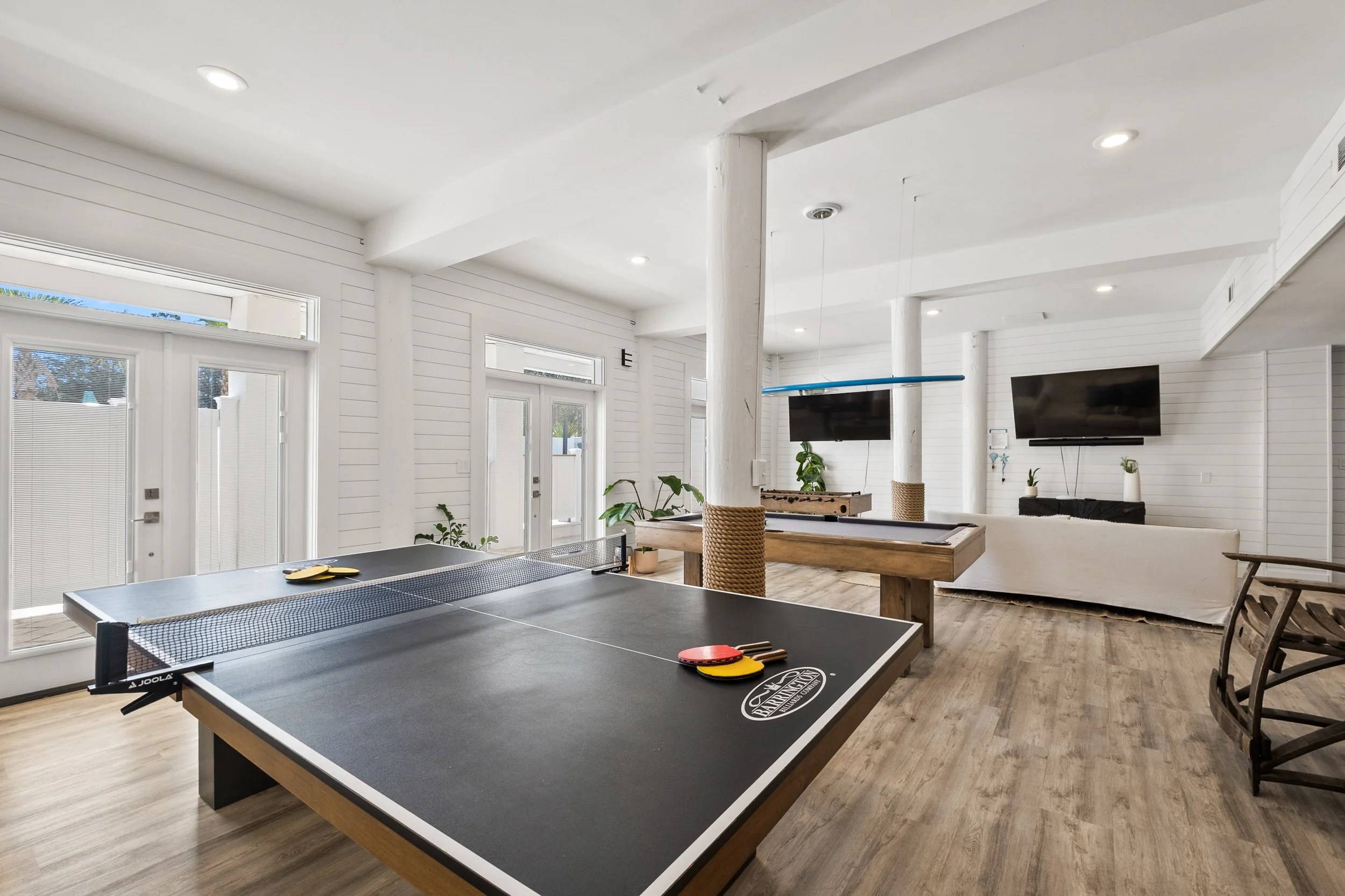 Interior of a recreational room featuring a ping pong table, foosball table, wall-mounted TV, sofa, and wood flooring with white walls and ceiling.