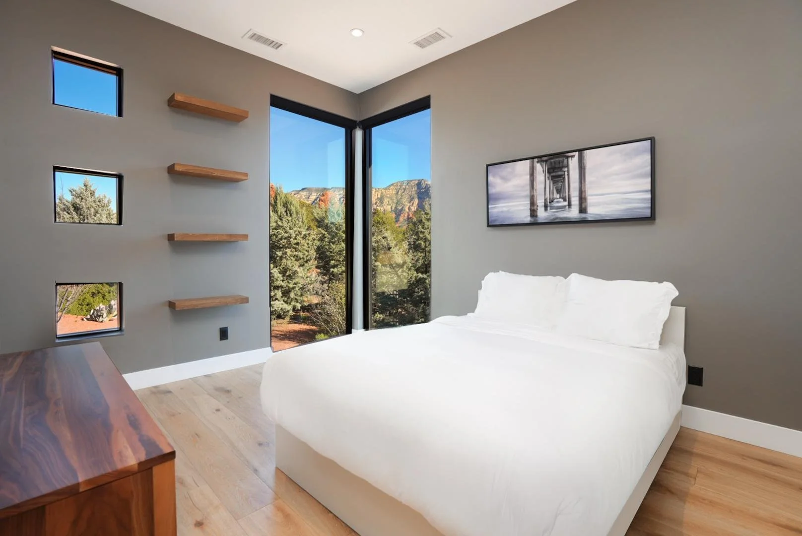 Modern bedroom with large corner window showing trees and mountains outside, white bed with pillows, wooden dresser, wall-mounted artwork of a pier, and three floating shelves on a gray wall.