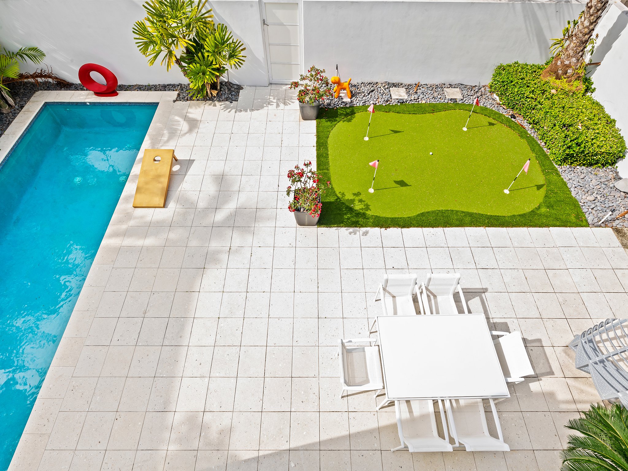 An aerial view of a backyard with a swimming pool, a small putting green with four flags, potted plants, a tree, and outdoor furniture.