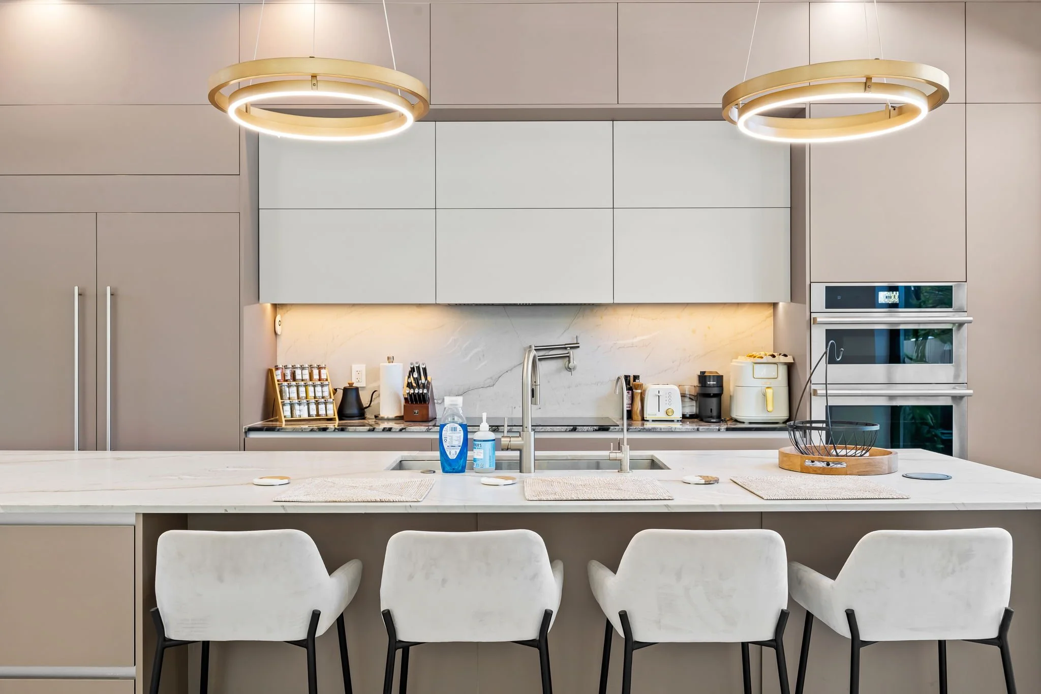 Modern kitchen with beige cabinetry, a large white island with bar stools, and built-in stainless steel oven and microwave. The kitchen features pendant lighting and various small appliances and decorative items on the countertops.
