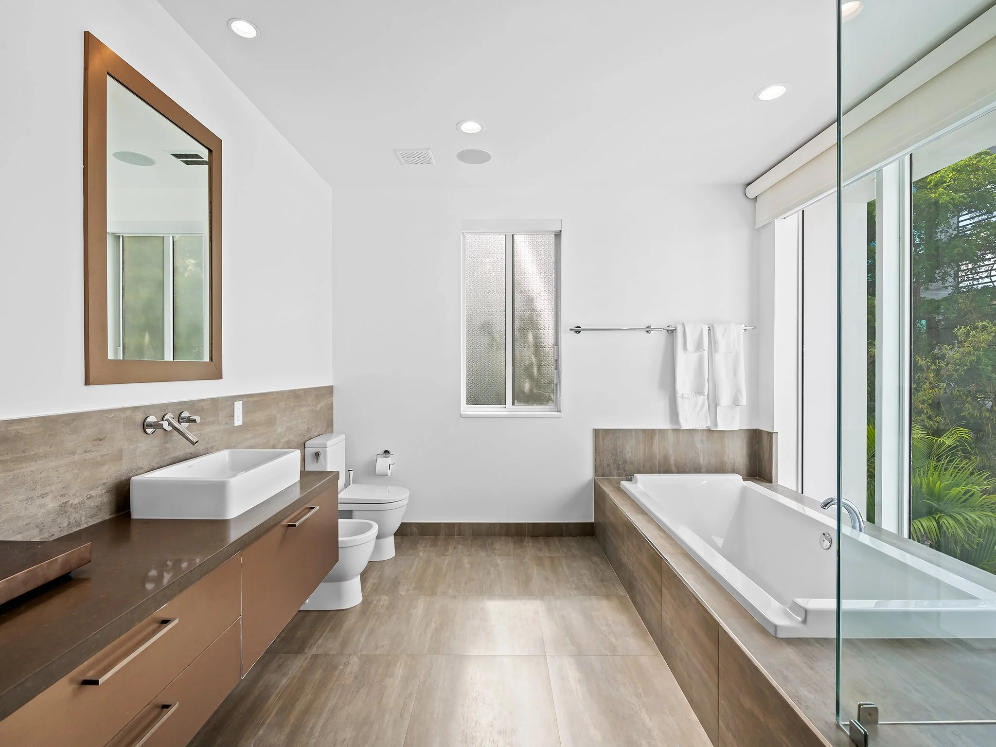 Modern bathroom with white walls, large window, wood-finished vanity, rectangular vessel sink, mirror, toilet, bathtub, and glass shower enclosure.