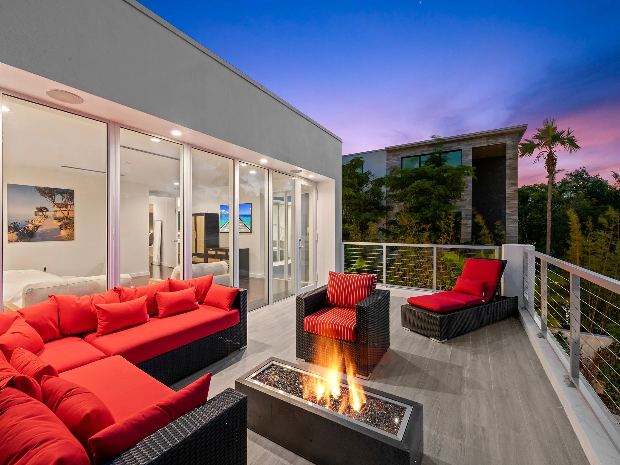 Modern outdoor balcony with red cushioned rattan furniture, a firepit table, and a view of a sunset sky with purple and pink hues, trees, and neighboring buildings.