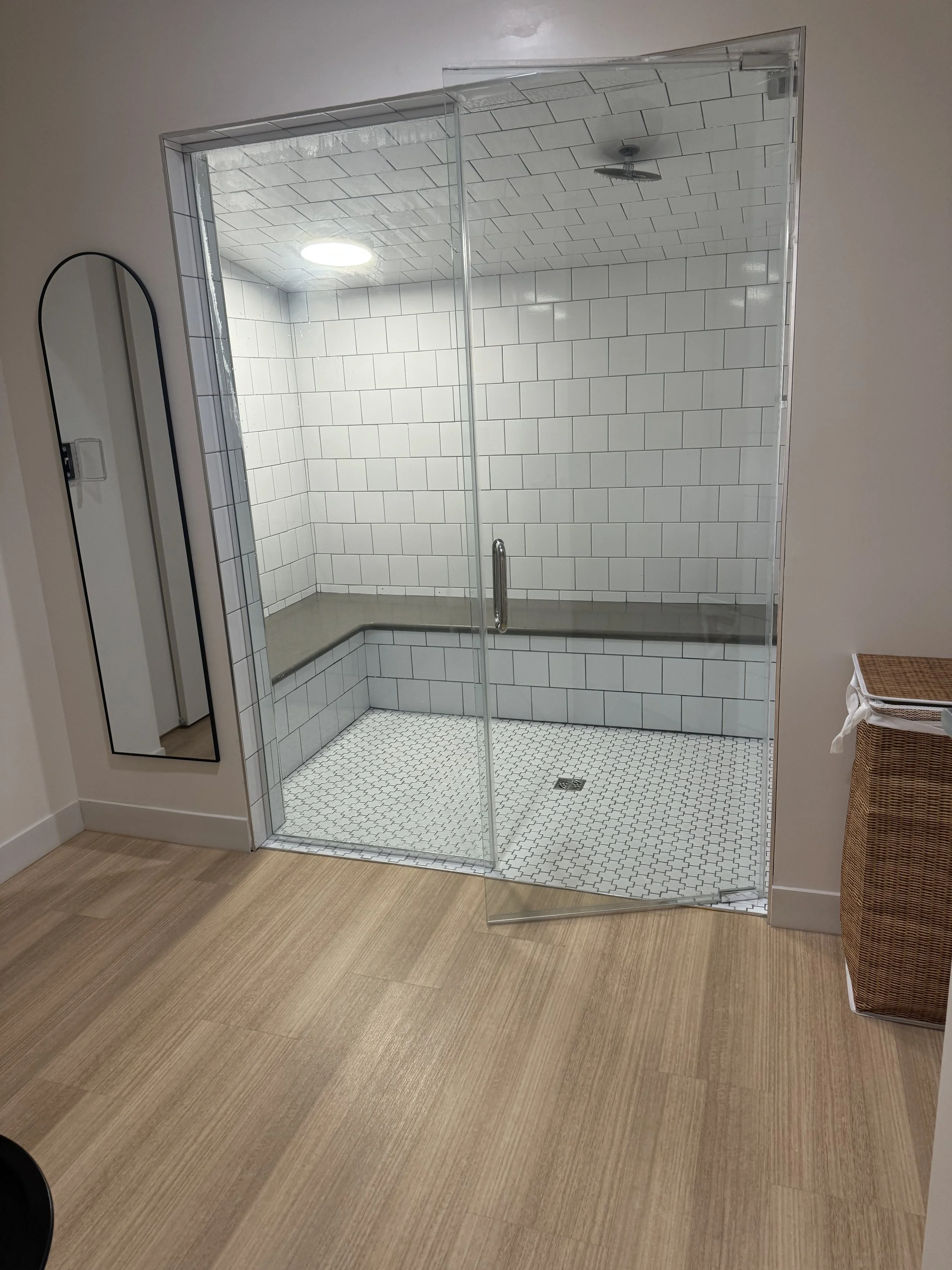 A minimalist shower with white subway tiles, a glass door, and built-in seat, located in a room with light wood flooring and a mirror on the wall.