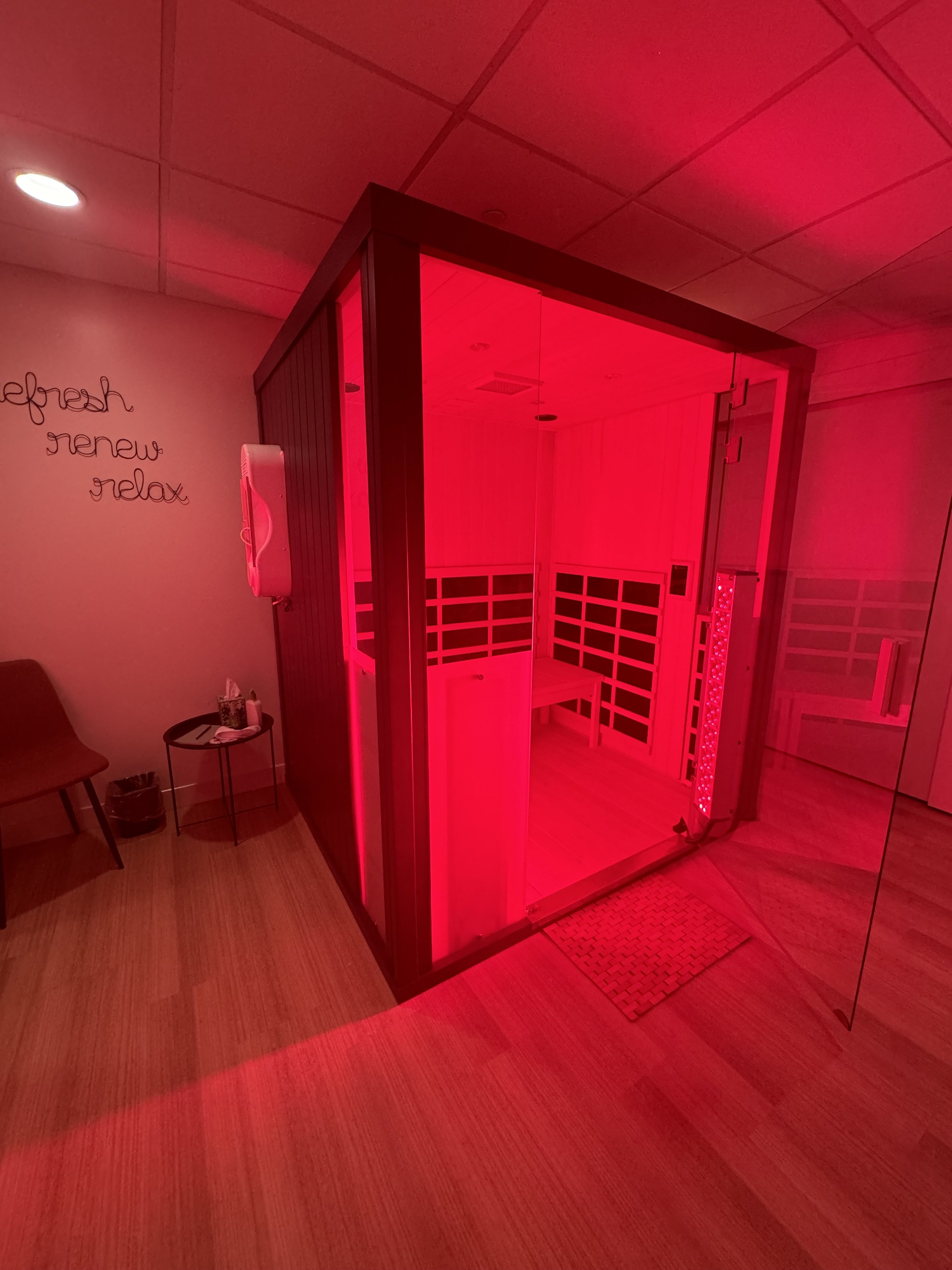 A red-lit sauna room with glass walls, inside wooden benches, and a red LED heater along the wall. To the side, there is a waiting area with a chair, a small table, a telephone, and a wall sign that reads 'refresh, renew, relax'.