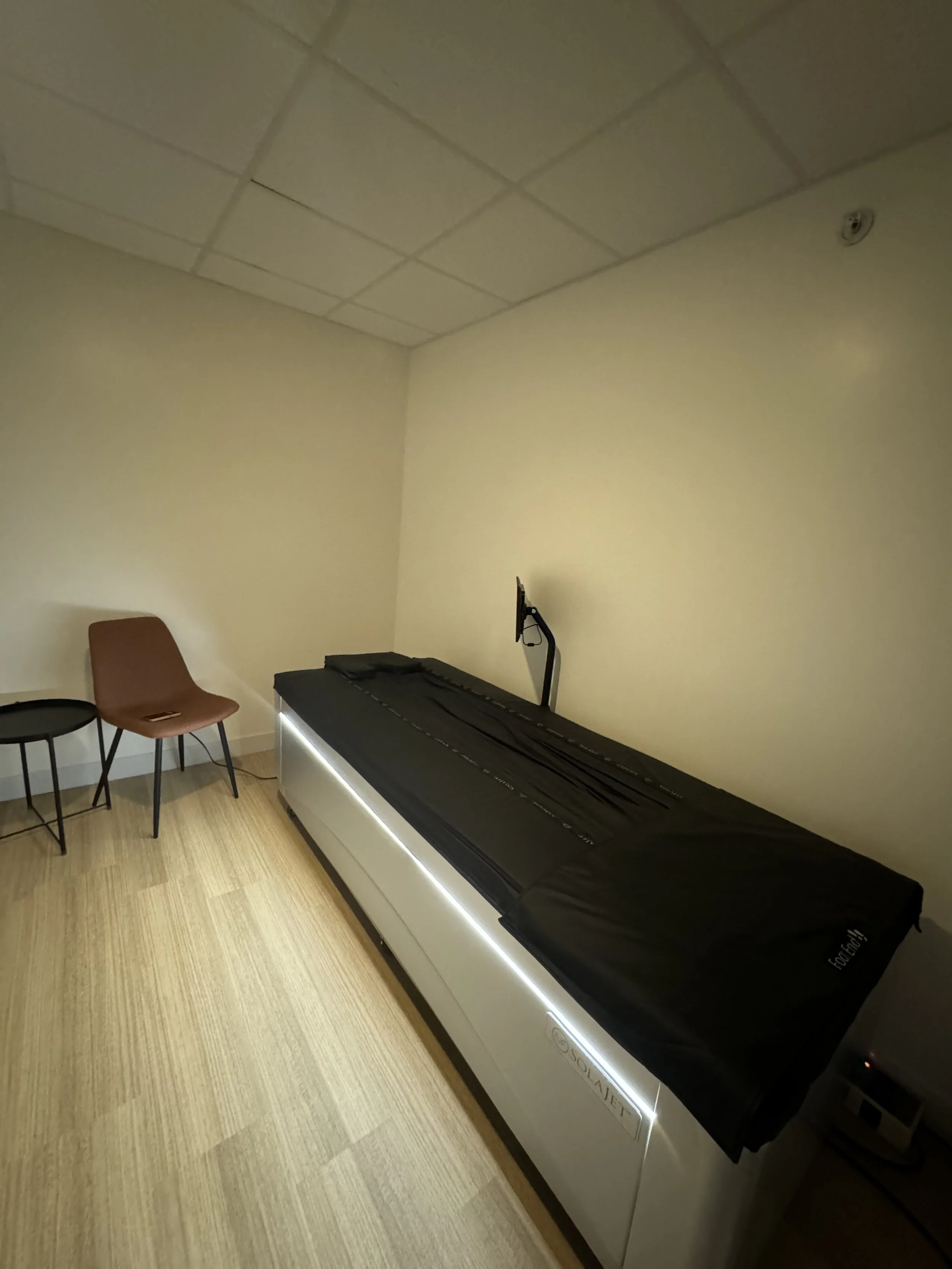 An empty medical or dental examination room with a bed covered in black sheet, a brown chair, and a small round black side table.