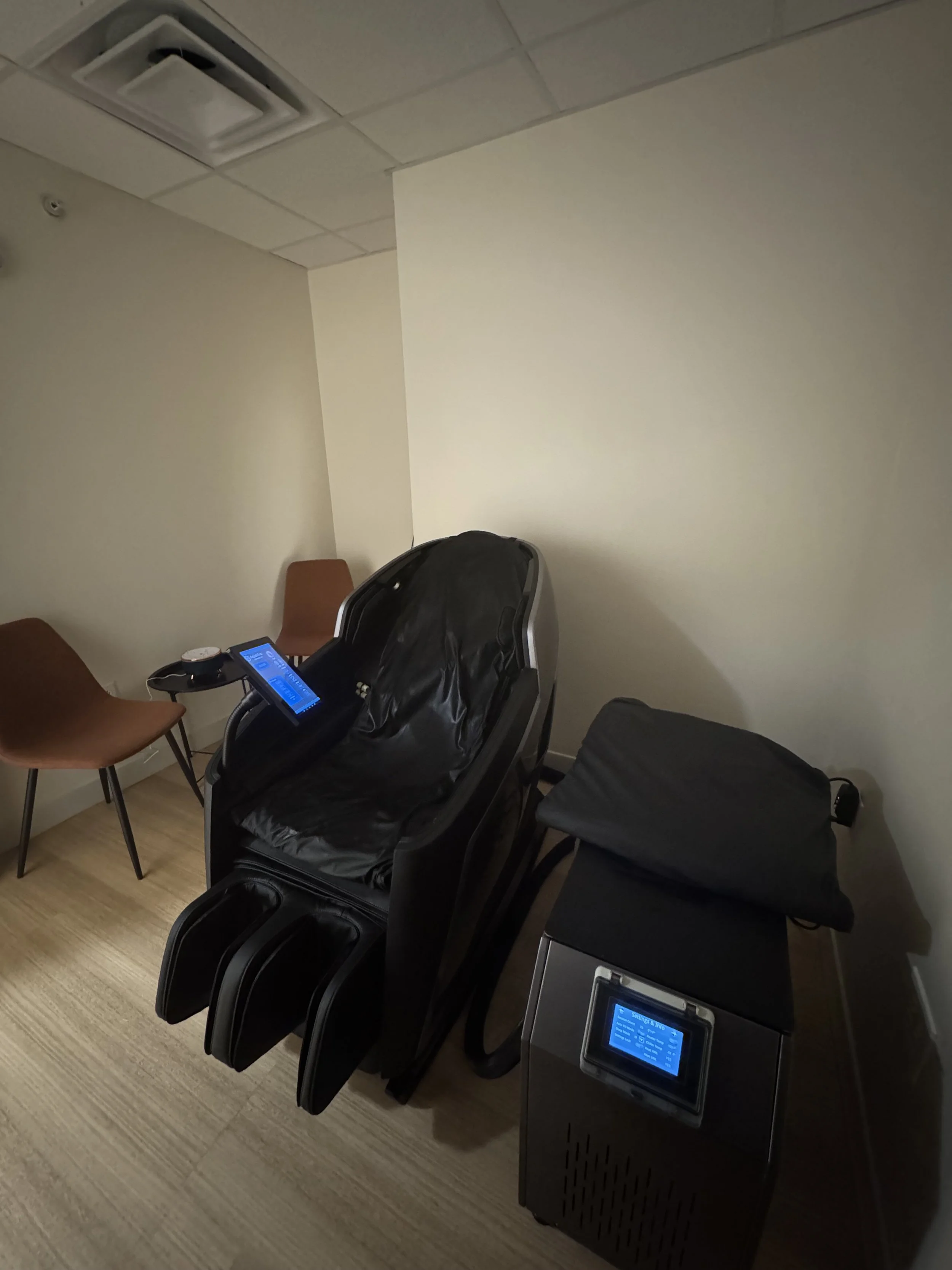 Relaxation chair with a control panel, two chairs, and a machine with a screen in a room with beige walls and wood flooring.