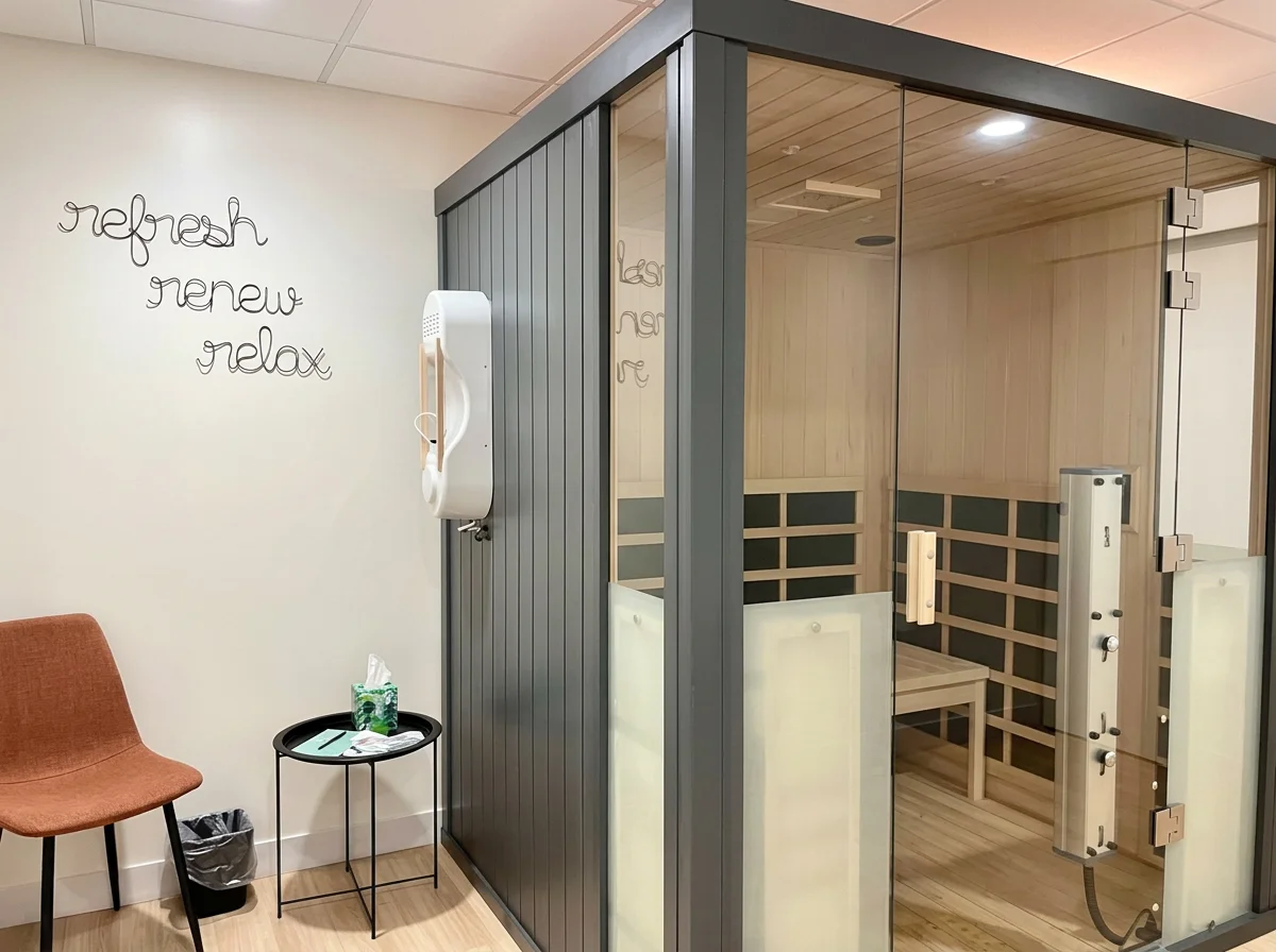 A modern infrared sauna with a glass door in a cozy health or wellness room. There is a wall with signed words 'refresh, renew, relax,' and a small chair with tissue box and wastebasket beside it.