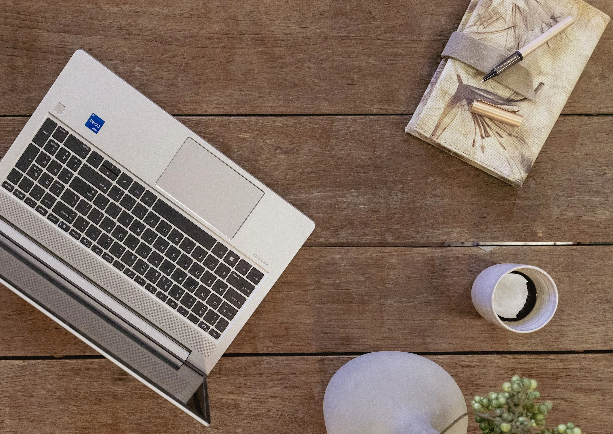 Een laptopschakeling op een houten tafel met een bloemstuk in een witte vaas, een schrift en pennen.