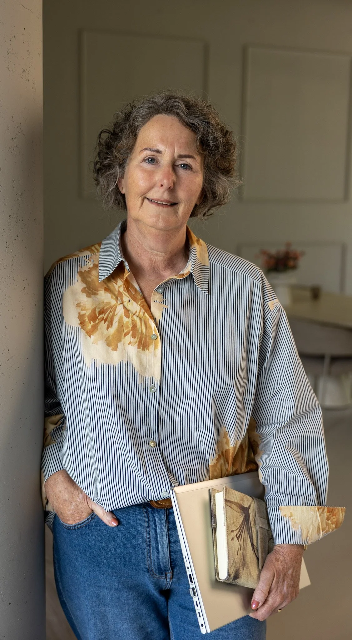 Een vrouw met kort, krullend grijs haar die half naast een muur staat. Ze draagt een blouse met een bloemmotief en verticale strepen, en blauwe jeans. Ze houdt een open laptop, een boek en een tablet in haar handen.