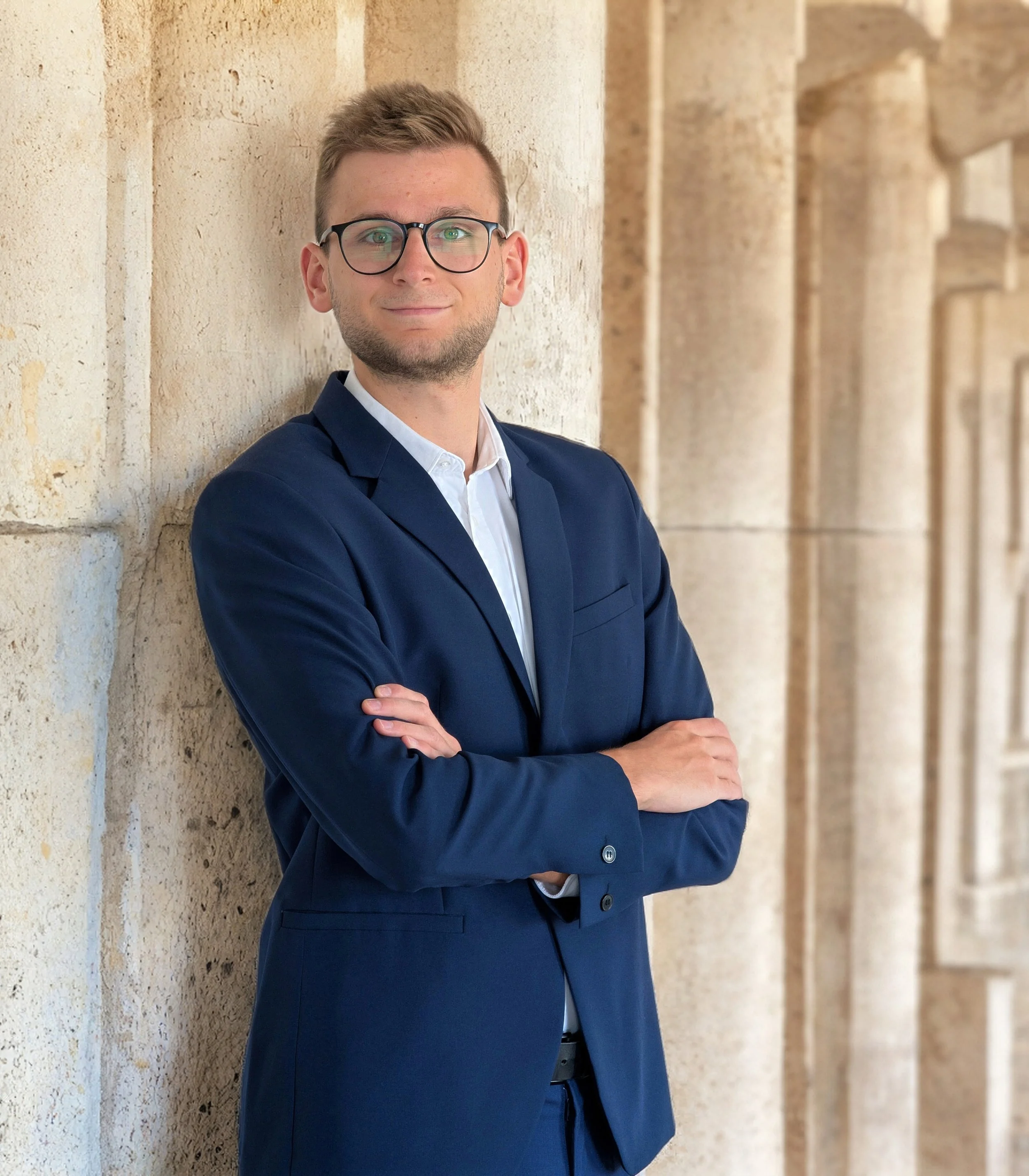 Ein junger Mann in einem blauen Anzug und weißen Hemd, trägt Brille und steht mit verschränkten Armen vor einer steinernen Wand, lächelt leicht.