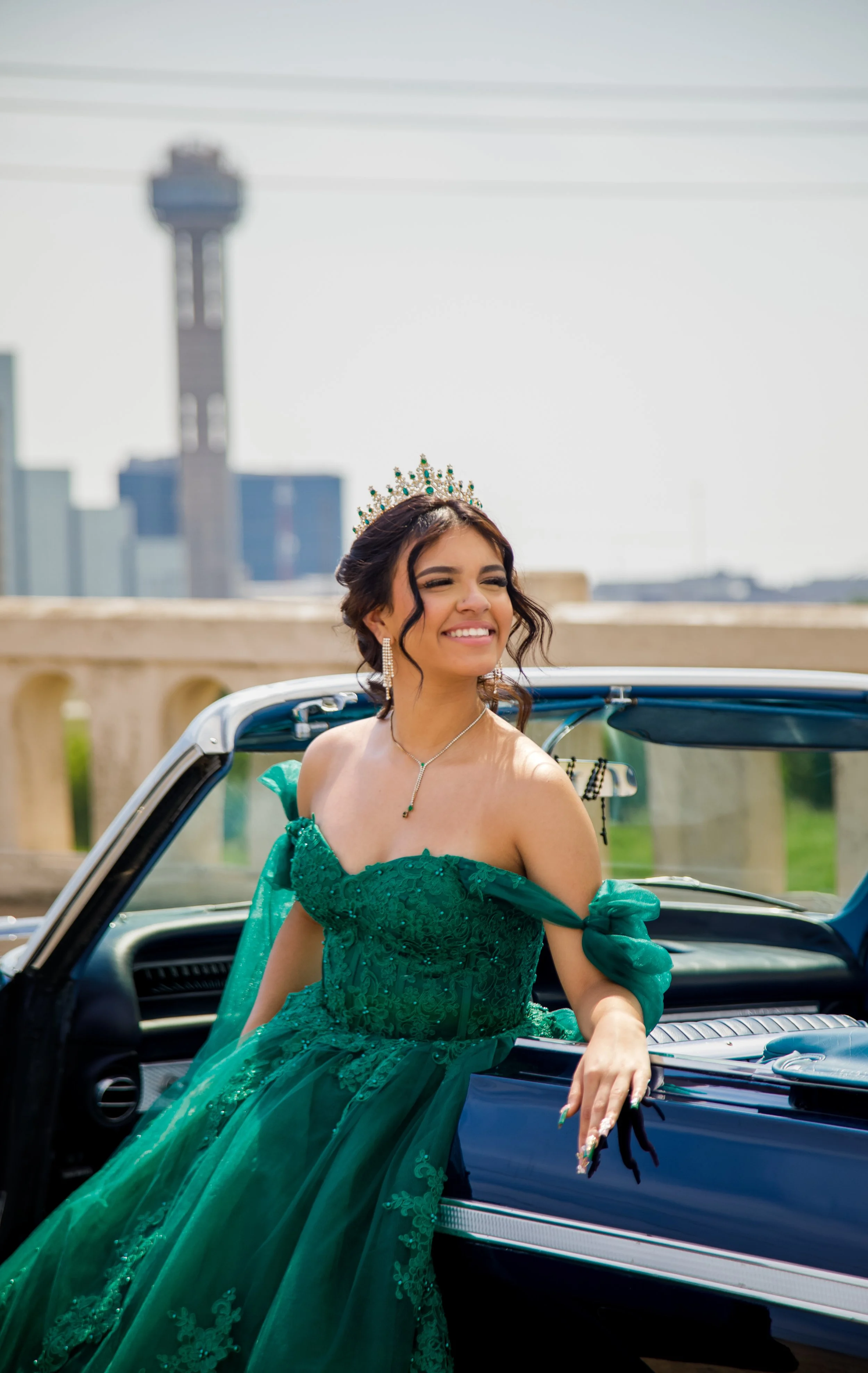 Young woman in a green gown and tiara, smiling, leaning on an open classic car, city skyline in the background.