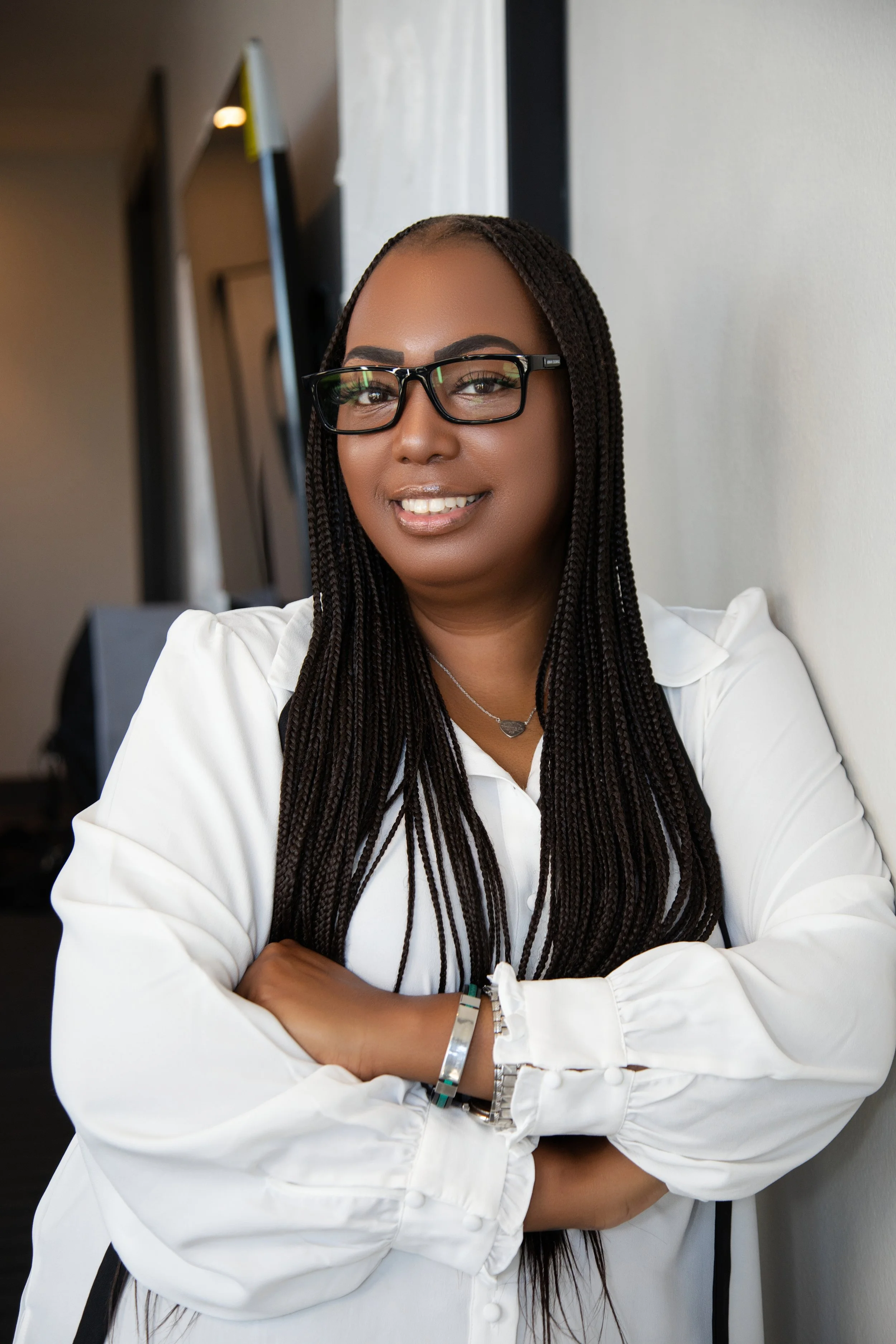 A woman with long braided hair, wearing glasses and a white blouse, smiling with arms crossed and leaning against a wall.