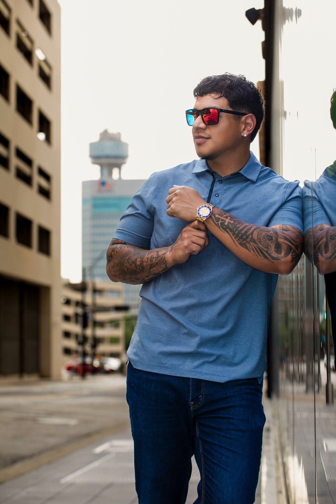A young man wearing a blue polo shirt, jeans, and sunglasses with red and blue lenses, standing outdoors by a reflective surface in an urban area. He has tattoos on his arms, a watch, and an earring, and is looking to the side with a confident expres