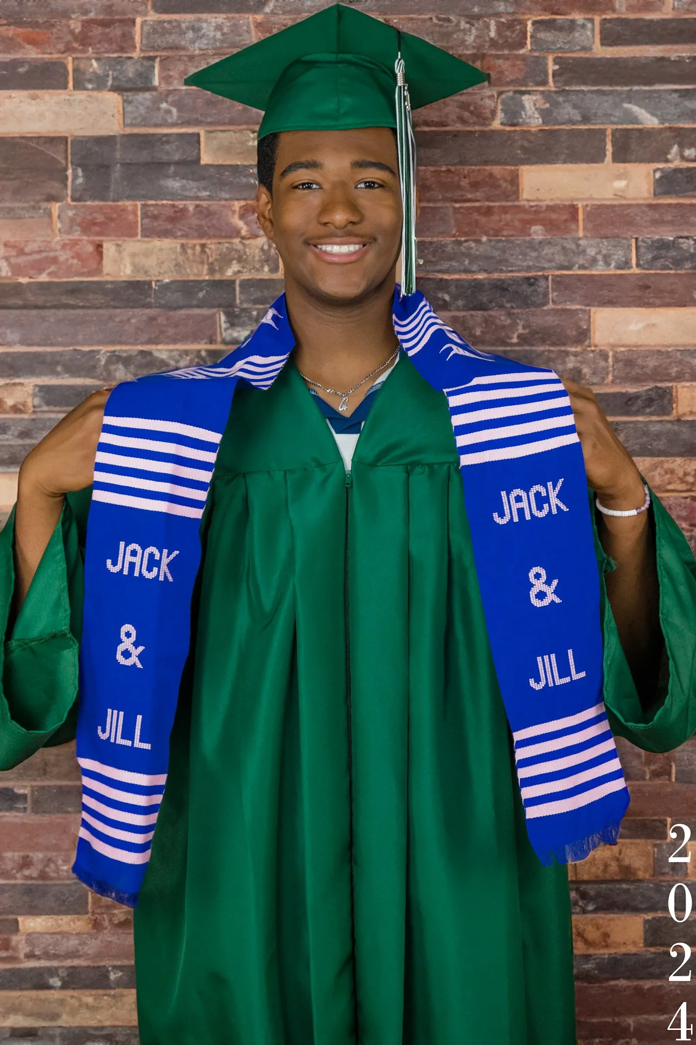 A young man in a green graduation cap and gown, holding a blue stole with white stripes and the text 'JACK & JILL'. He is smiling, standing in front of a brick wall, with the year '2024' written vertically in the lower right corner of the image.