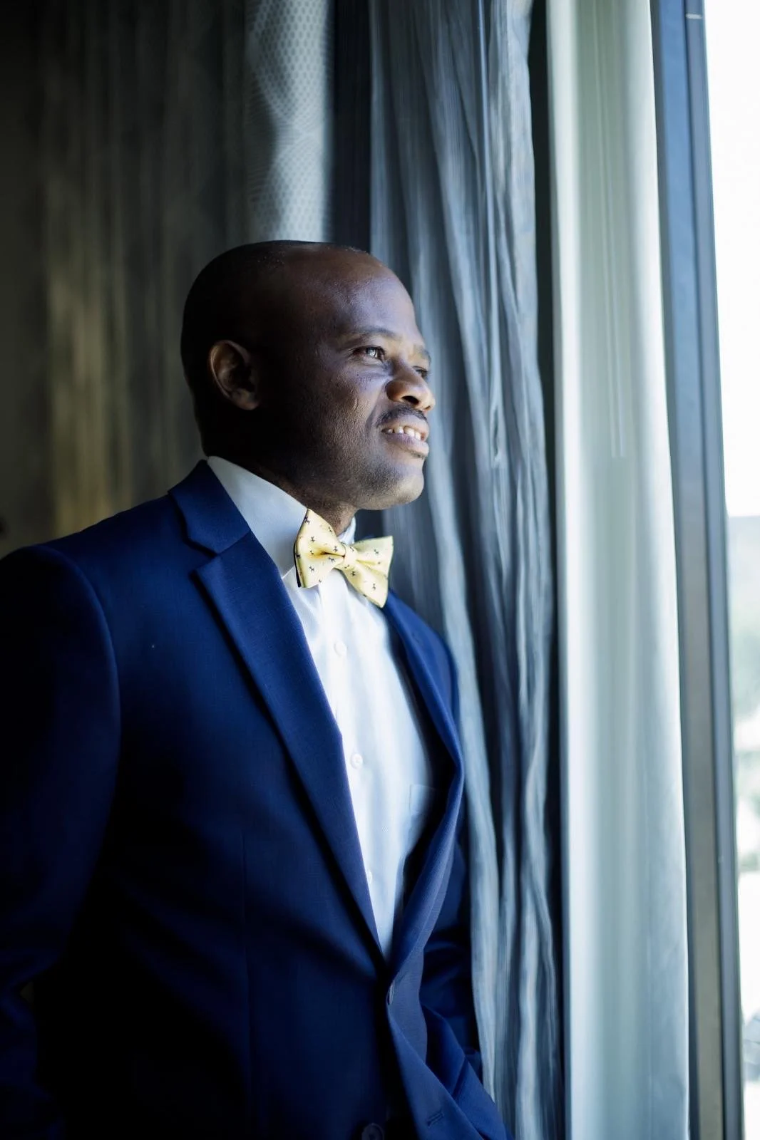 Man in a navy blue suit, white shirt, and yellow bow tie looking out the window.