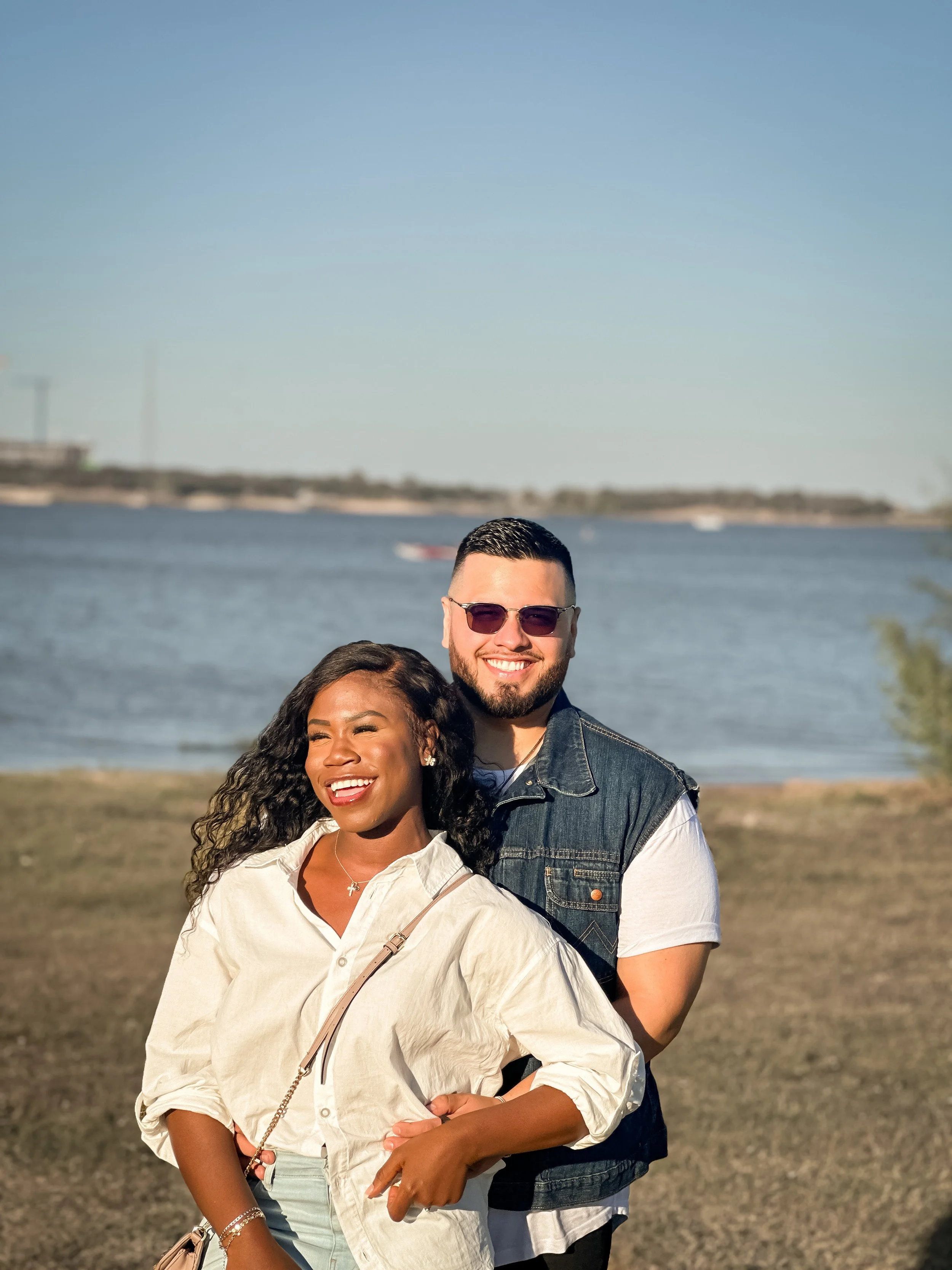 A smiling woman and man stand outdoors near a body of water on a sunny day. The woman has curly hair, is wearing a light-colored shirt, and the man has short dark hair, a beard, sunglasses, and a denim vest over a white t-shirt.
