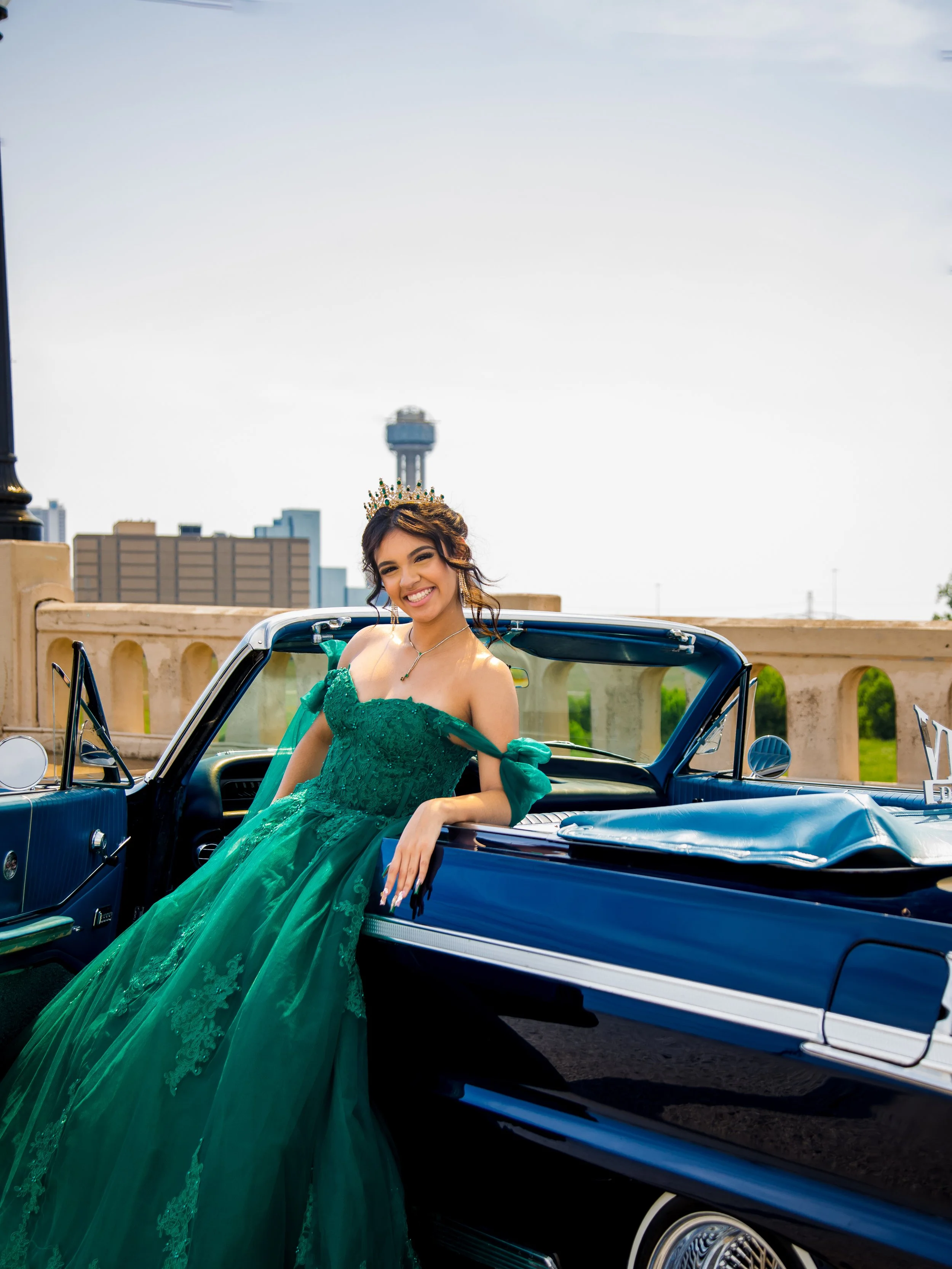 A woman in an emerald green gown and tiara leaning on a vintage blue convertible car, smiling with city skyline in the background.