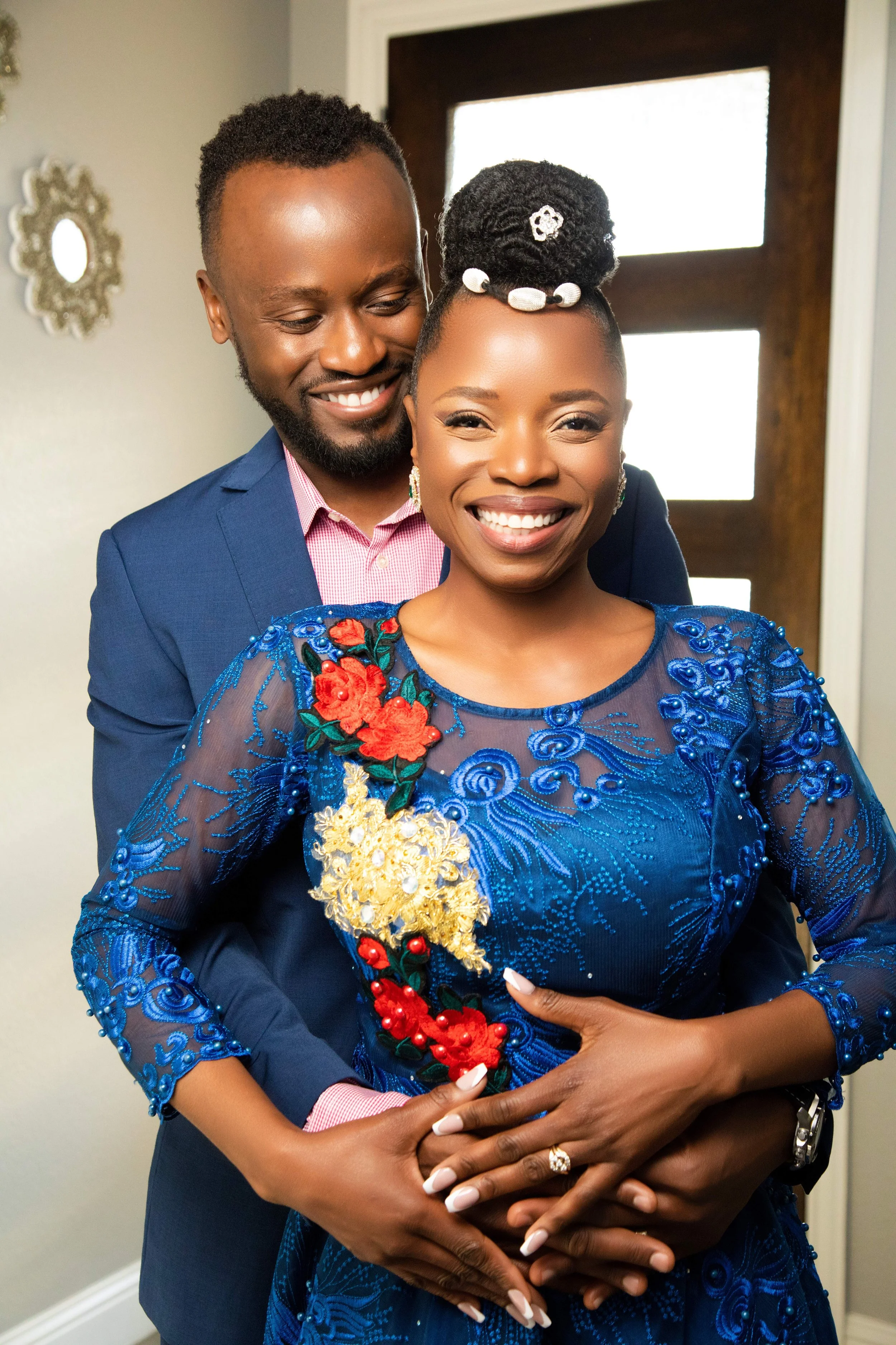 A smiling woman with an updo hairstyle and a blue embroidered dress, being embraced from behind by a smiling man in a blue suit, standing in a warmly lit room by a window.
