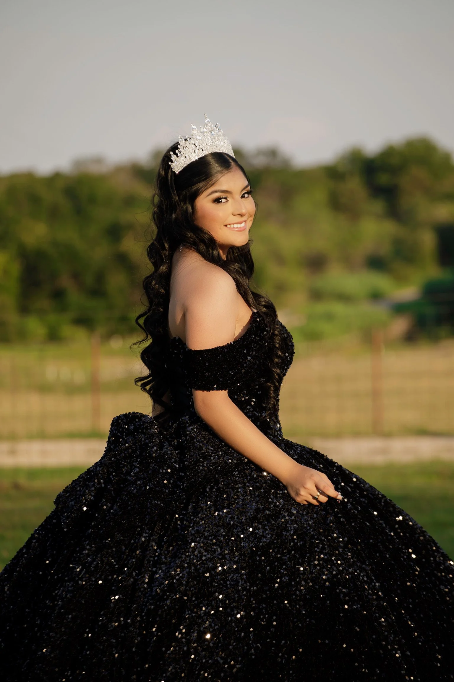 young girl with a tiara, in a black sparkly dress, outside near trees, at sunset