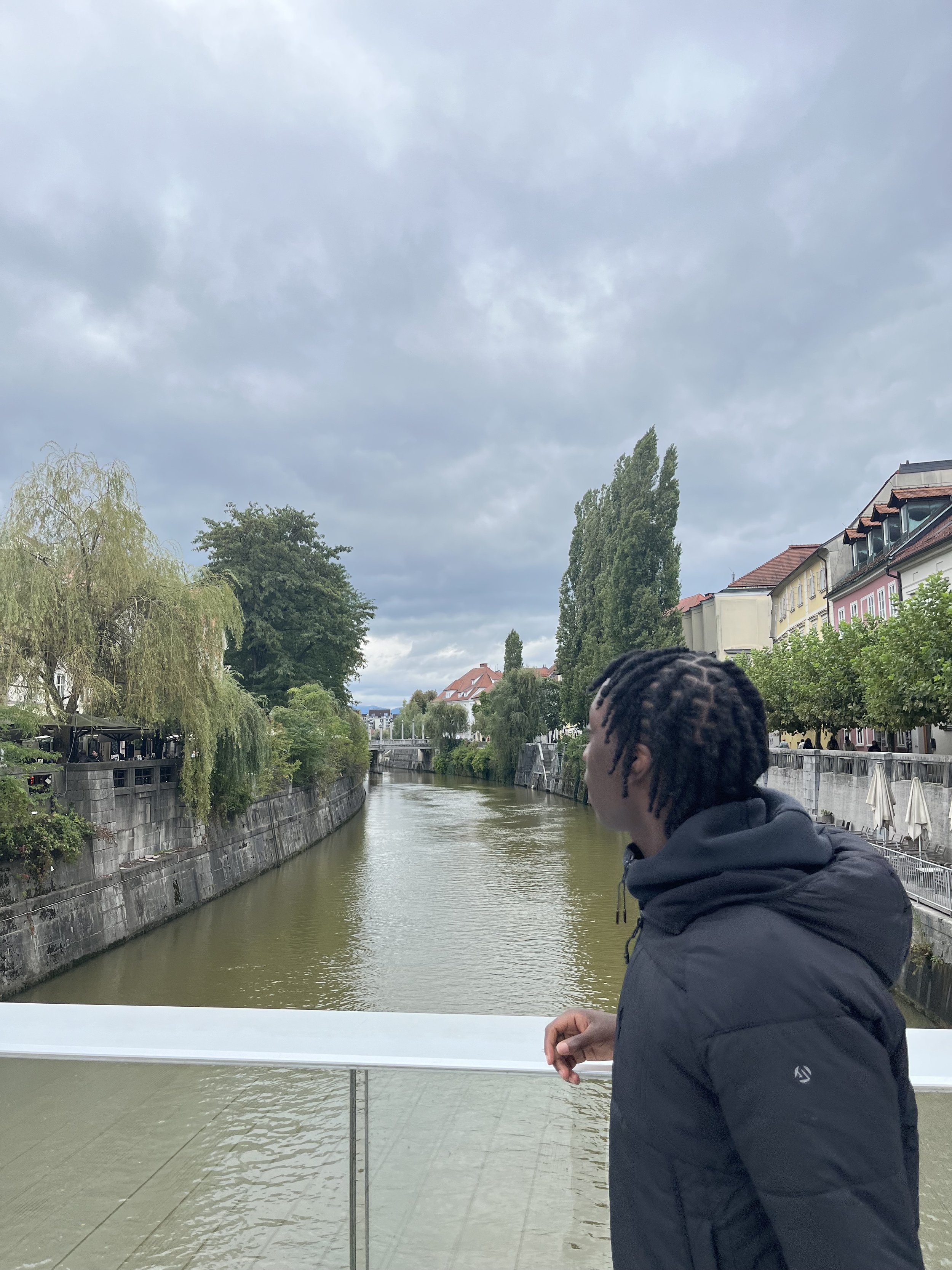 Junge Person mit Dreadlocks in dunkler Jacke steht an einer Brüstung und schaut auf einen Fluss in einer Stadt mit Bäumen und bunten Häusern bei bewölktem Himmel.