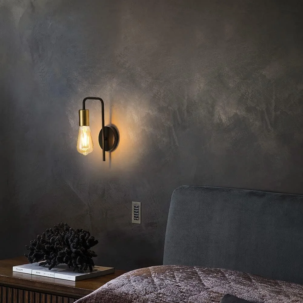A bedroom corner with a dark wall, modern wall sconce with exposed filament bulb, grey upholstered headboard, woven decor item on a wooden nightstand, and a book.