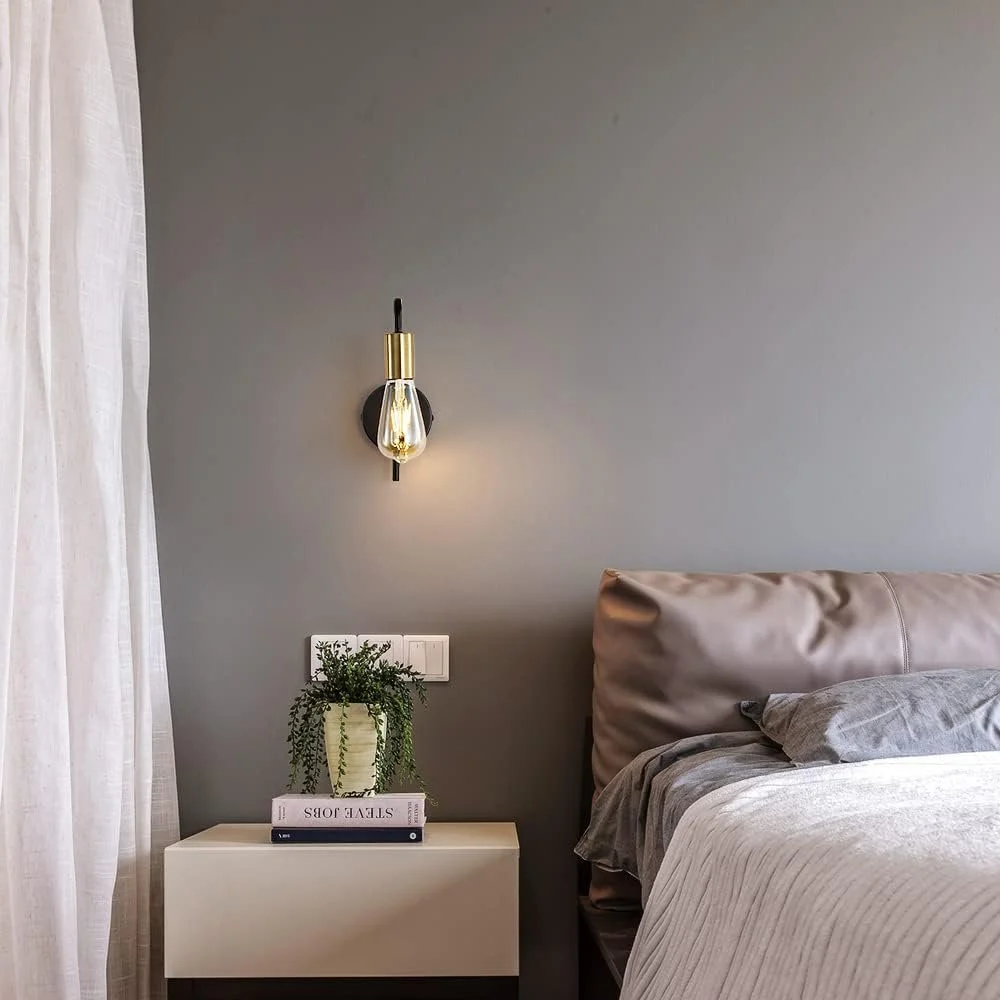 Contemporary bedroom with a beige upholstered headboard, gray bedding, a white nightstand, a green potted plant, a stack of books, a black wall-mounted light fixture, and white curtains on the left side.