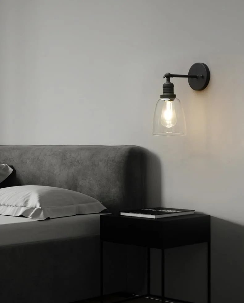 A bedroom with a gray upholstered bed, white pillow, black nightstand with books, and a wall-mounted black metal and glass light fixture.