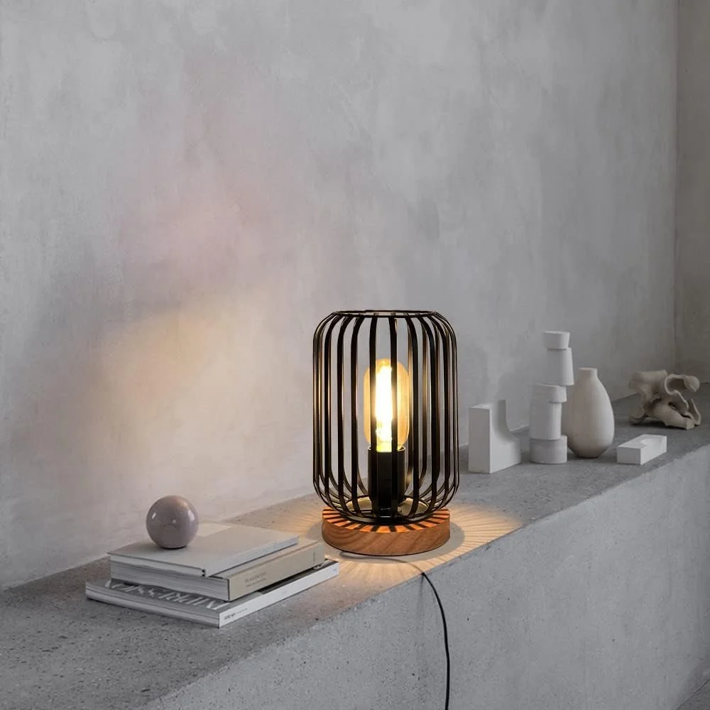 A modern black wireframe lamp with a wooden base illuminated on a concrete surface, surrounded by white decorative objects and books against a textured white wall.