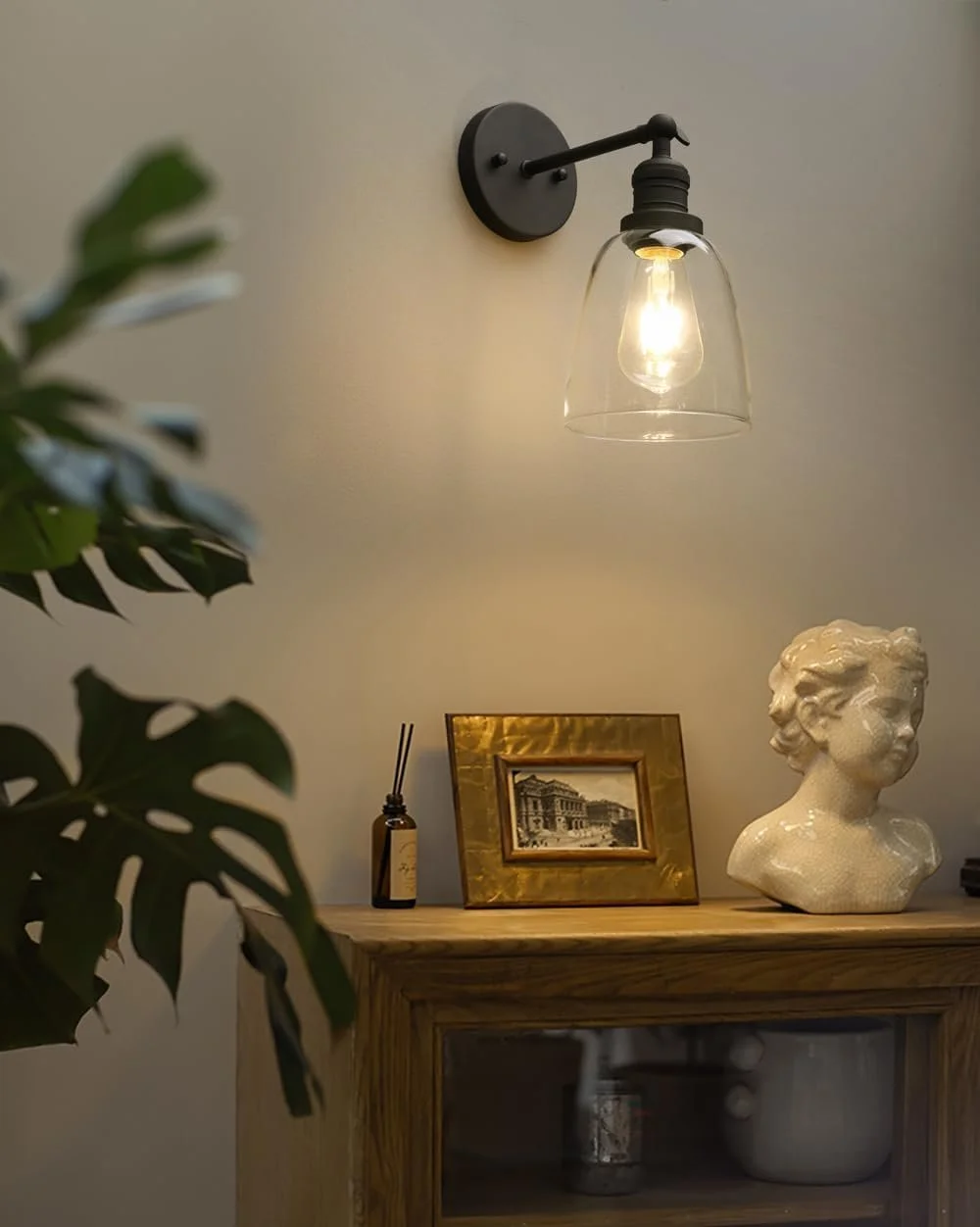 Interior scene with a wall-mounted black metal and glass wall sconce illuminating a corner, a wooden cabinet with a bust sculpture of a child's head, a framed photograph, and a small bottle with reed sticks, with a leafy green plant partially visible