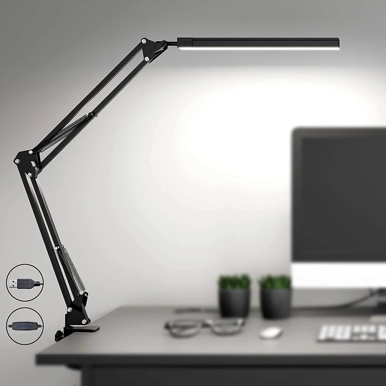 A modern black adjustable desk lamp on a grey desk with two small potted plants, a computer monitor, a keyboard, a mouse, and eyeglasses. The background is a plain light grey wall.