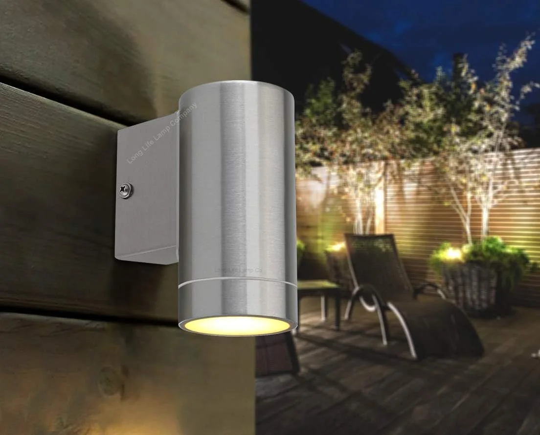 Close-up of a modern outdoor wall-mounted light fixture at night illuminating a backyard with patio chairs and potted trees.