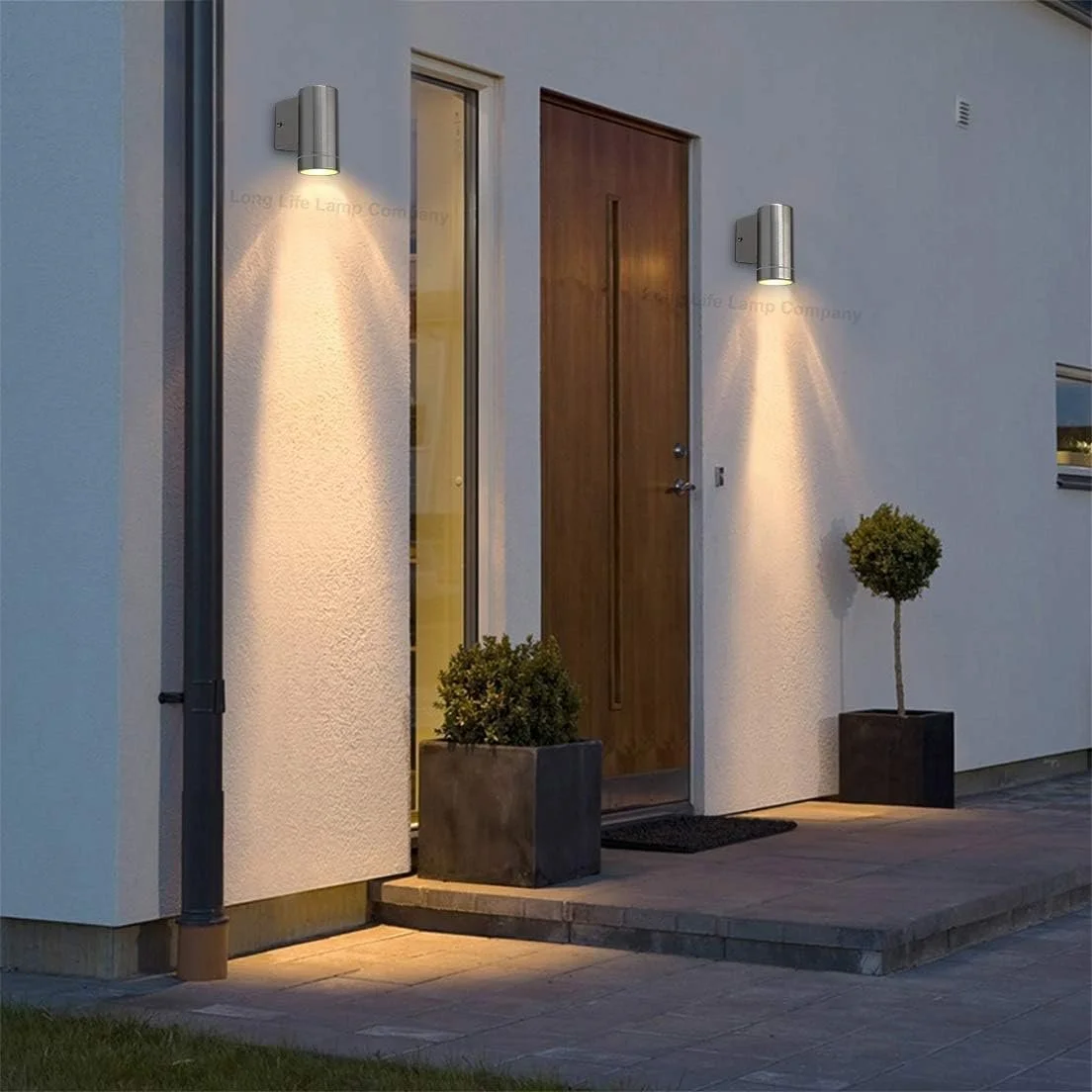 Exterior of a modern house entrance with two wall-mounted lights illuminating the doorway, a wooden door, two potted plants, and a small porch area at dusk.