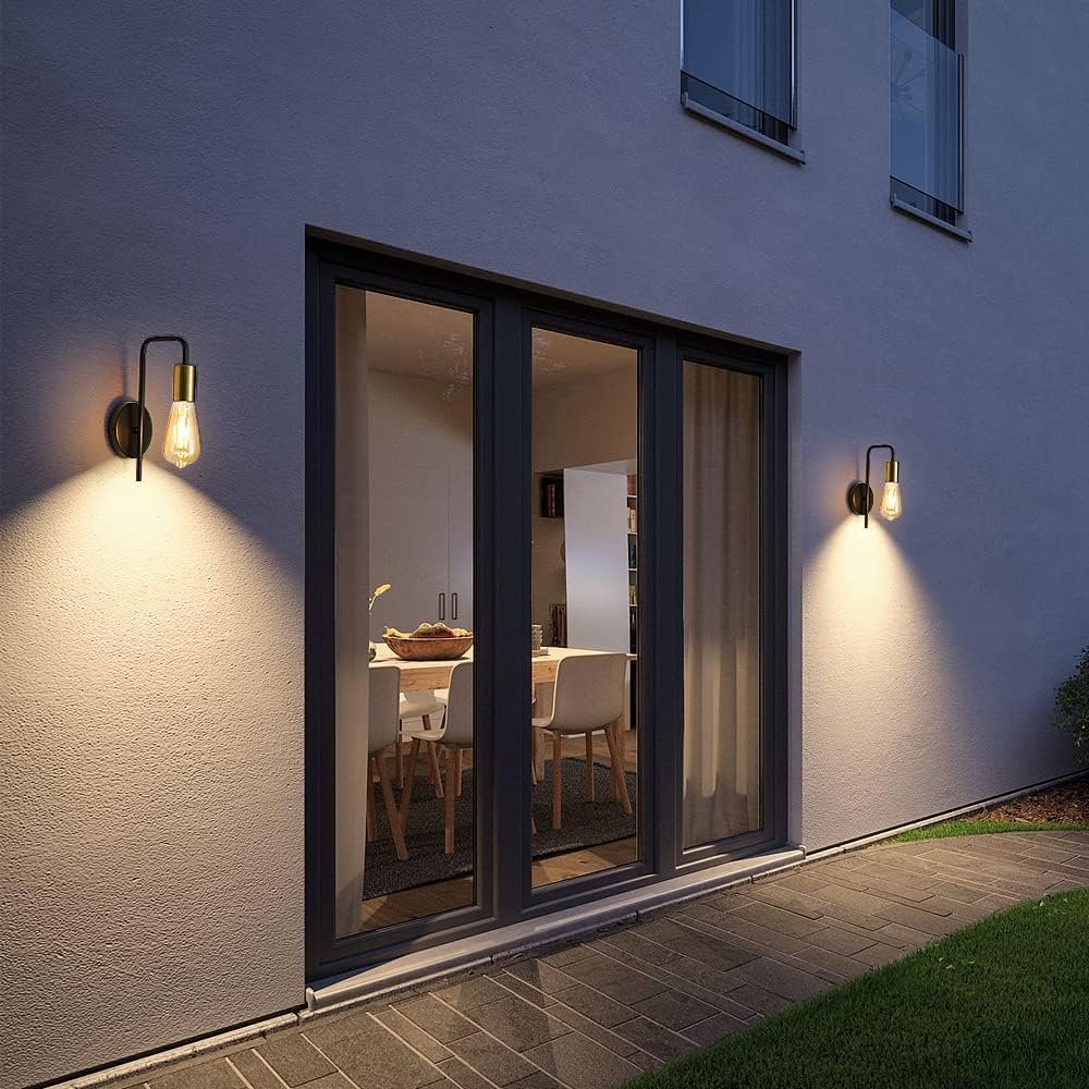 Exterior view of a modern house with large glass sliding doors showing a dining area inside, lit by wall-mounted outdoor lights, during evening.