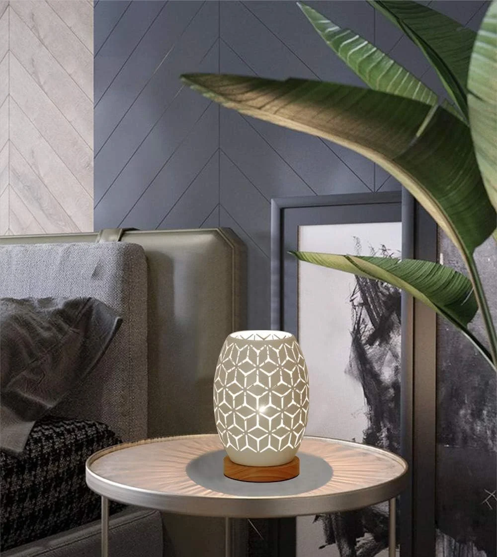 A modern living room corner featuring a stool with a table lamp on top. The lamp has a geometric cutout pattern emitting warm light. Large green leaves partially frame the scene, with a framed black and white artwork in the background, and a sofa wit