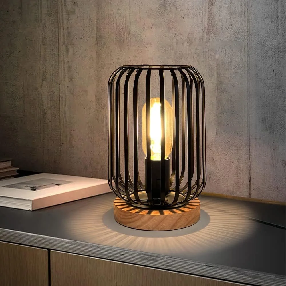 A table lamp with a wooden circular base, black metal cage-like frame, and an illuminated Edison-style filament bulb.