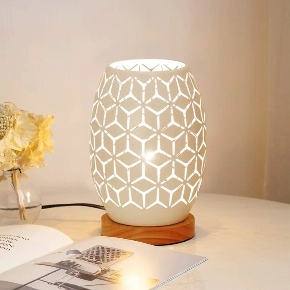 A white ceramic table lamp with a geometric cut-out pattern on a wooden base, glowing softly on a white table. There is a small vase with light-colored flowers and an open magazine nearby.