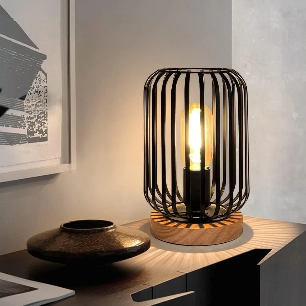 A modern table lamp with black vertical bars surrounding an Edison bulb, sitting on a wooden base, on a dark-colored table with a decorative brown bowl nearby.