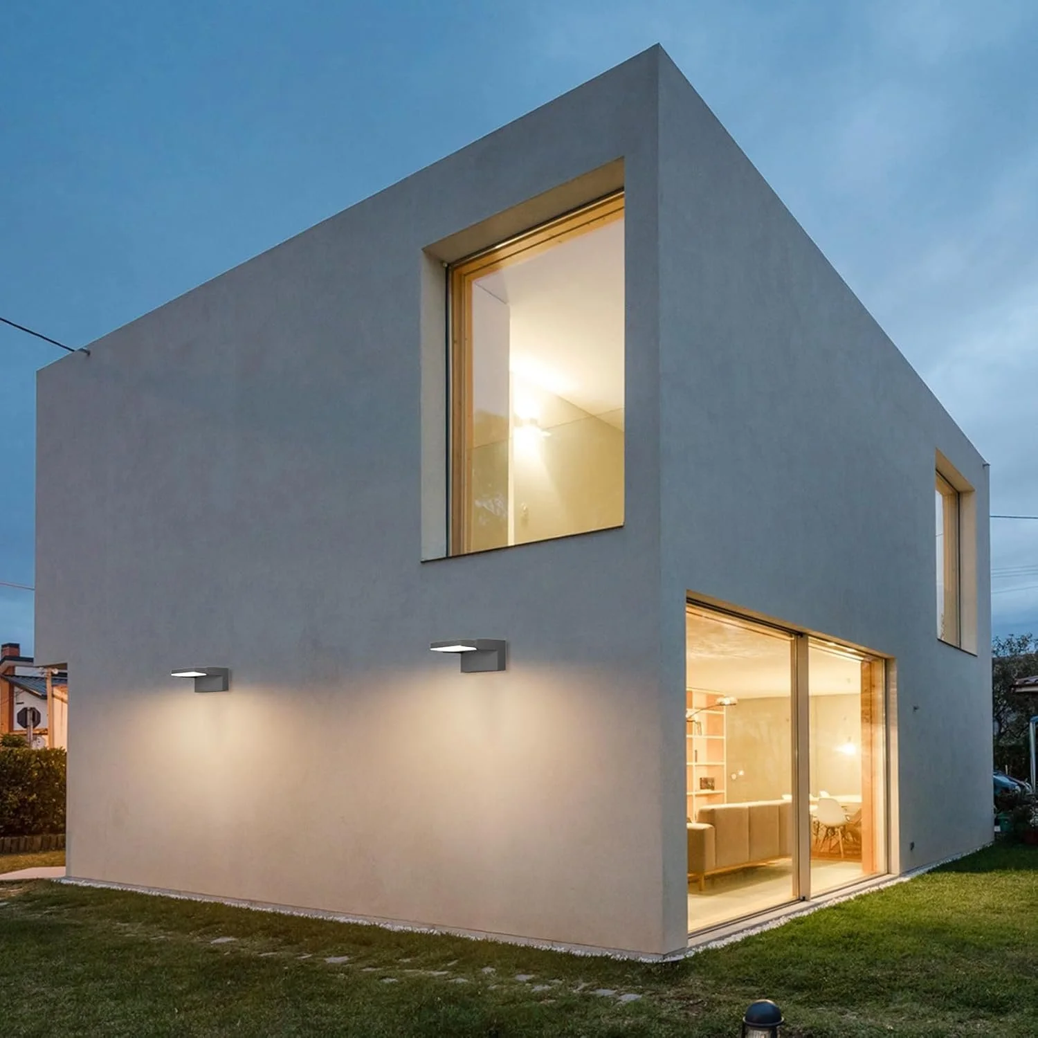 Modern two-story white house with large windows at dusk, interior lights on, and outdoor grass area.